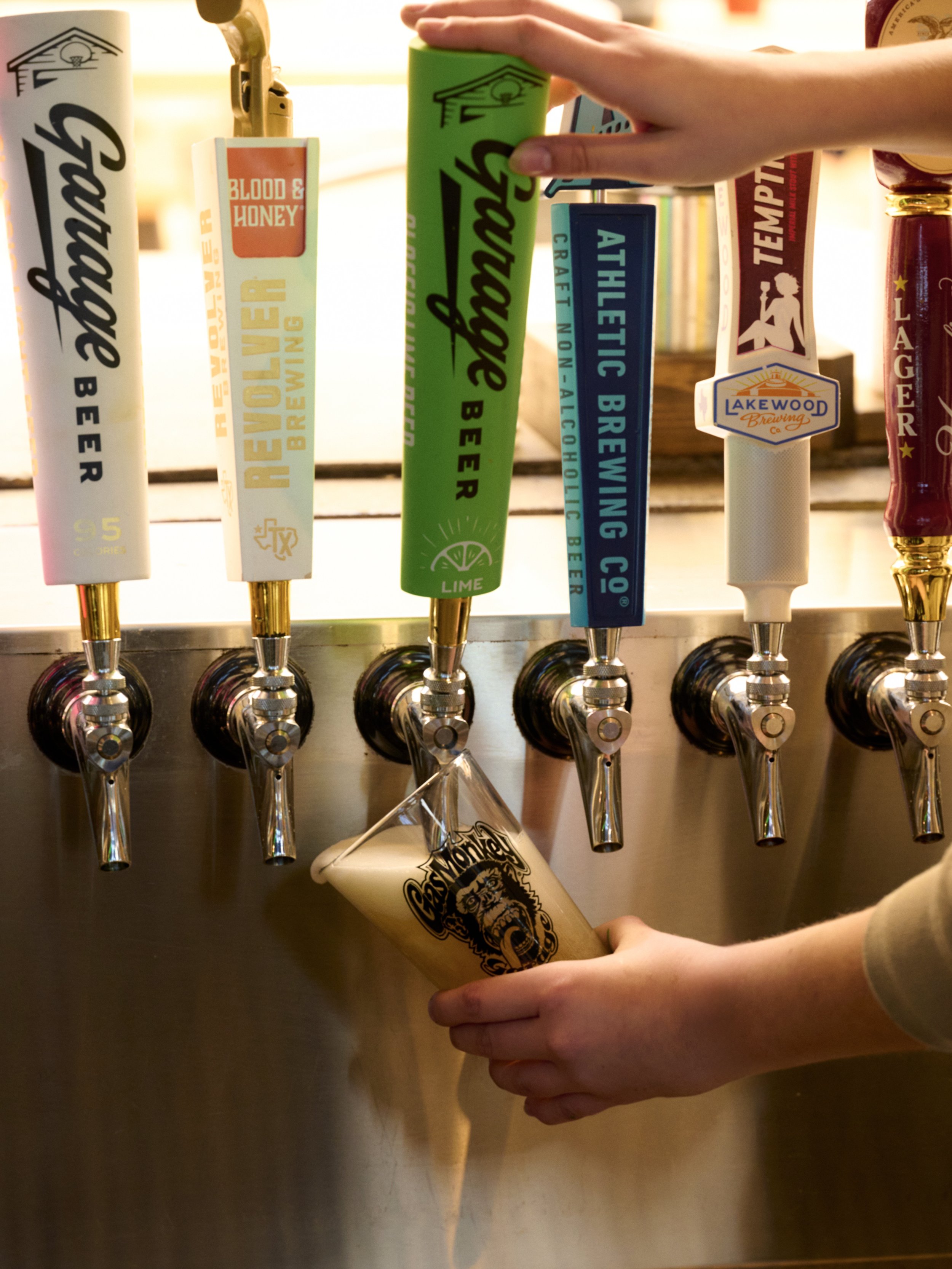 A person fills a glass Gas Monkey Garage pint with draft beer from a beer tap at a bar or brewery. The tap handles are decorated with various colorful labels, including 'Gooey' and 'Blood & Honey' from Lakewood Brewing.