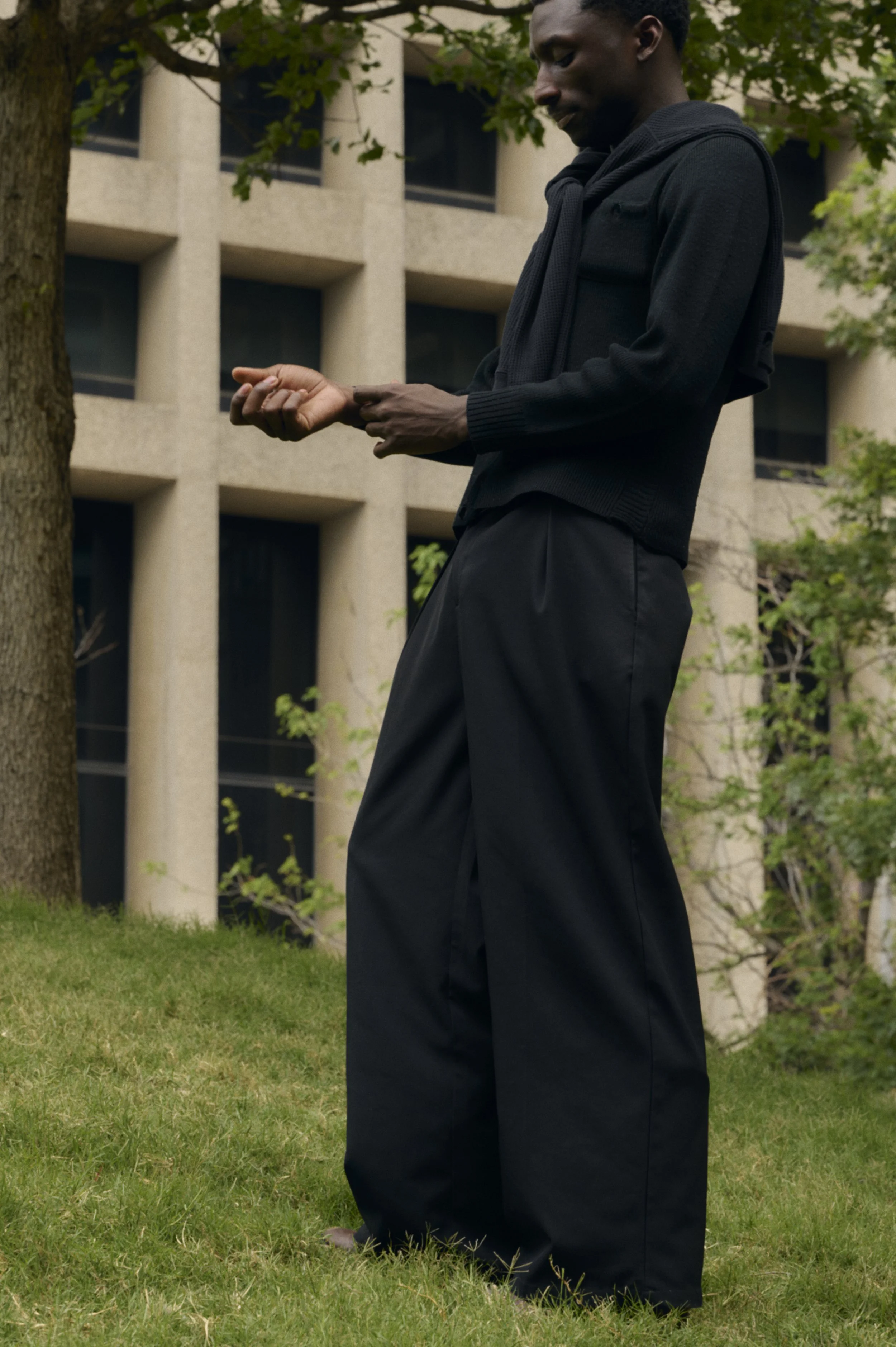 A man dressed in black, standing on a grassy area outdoors, looking at his wristwatch, with a building and trees in the background.