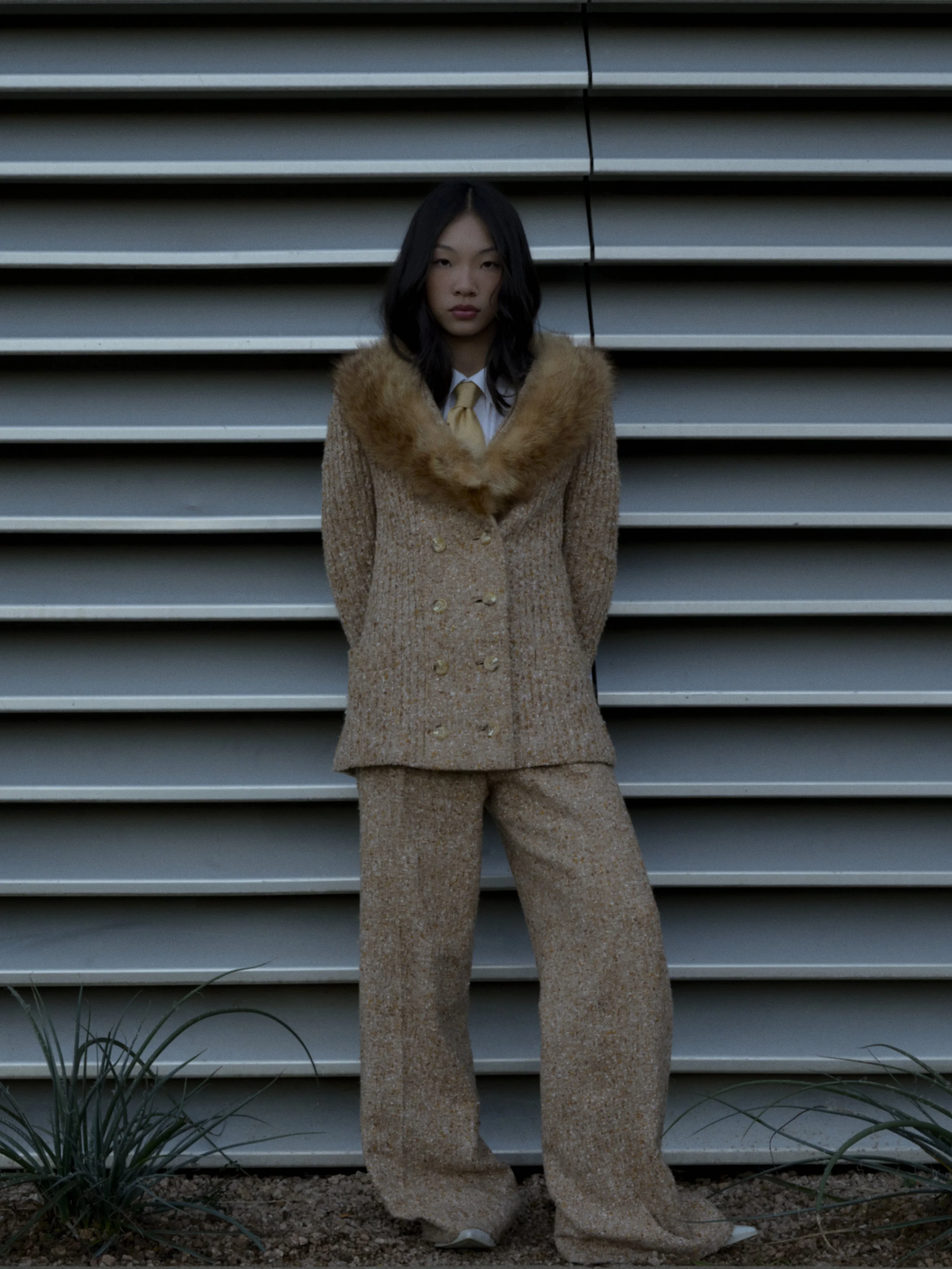 Young woman with black hair, wearing a beige coat with a fur collar and matching beige pants, standing in front of metal stairs.
