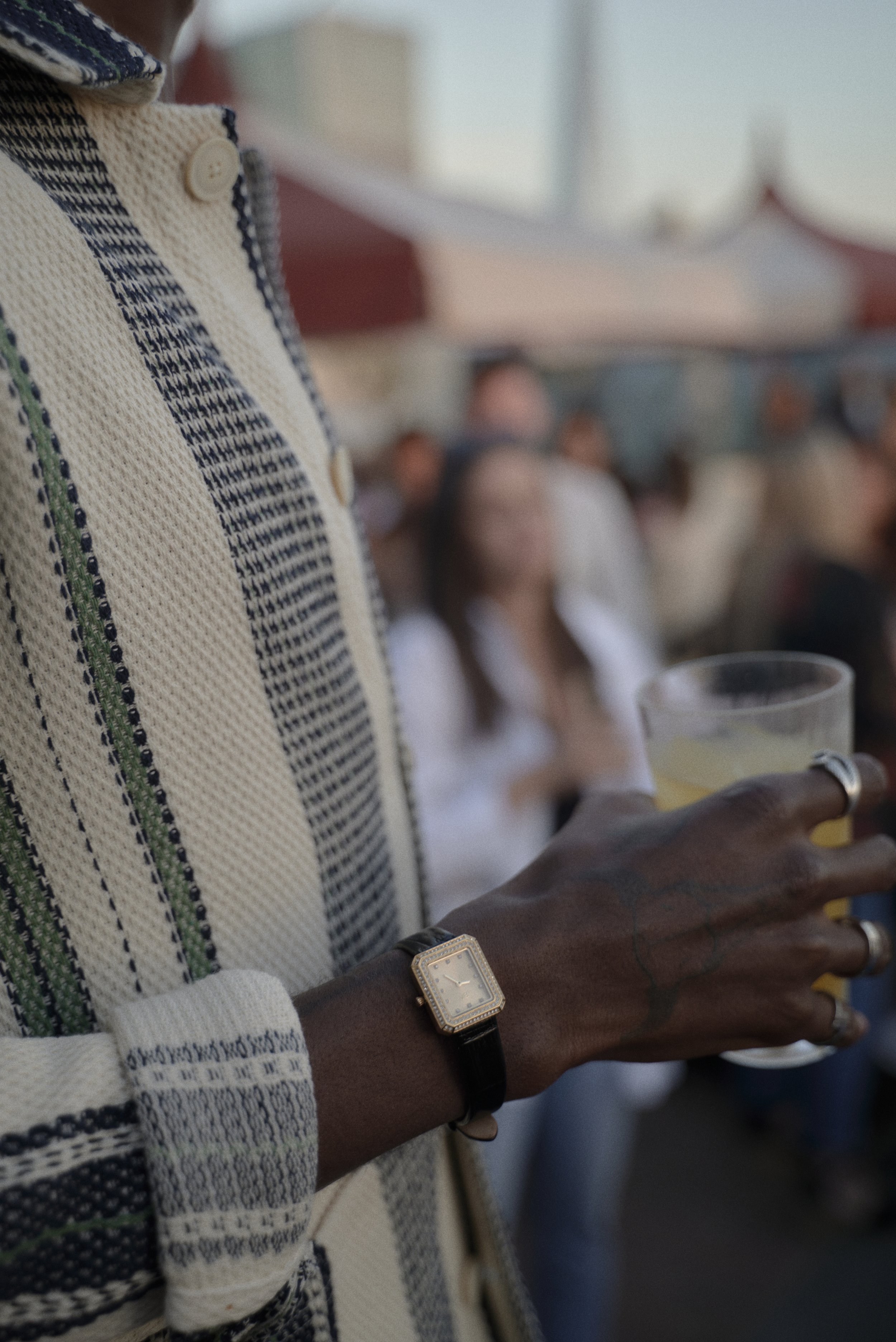 Close-up of a person's hand holding a clear plastic cup filled with a yellow beverage, wearing a square-faced watch with a black strap and a rectangular face, in a crowded outdoor setting with blurred people in the background.