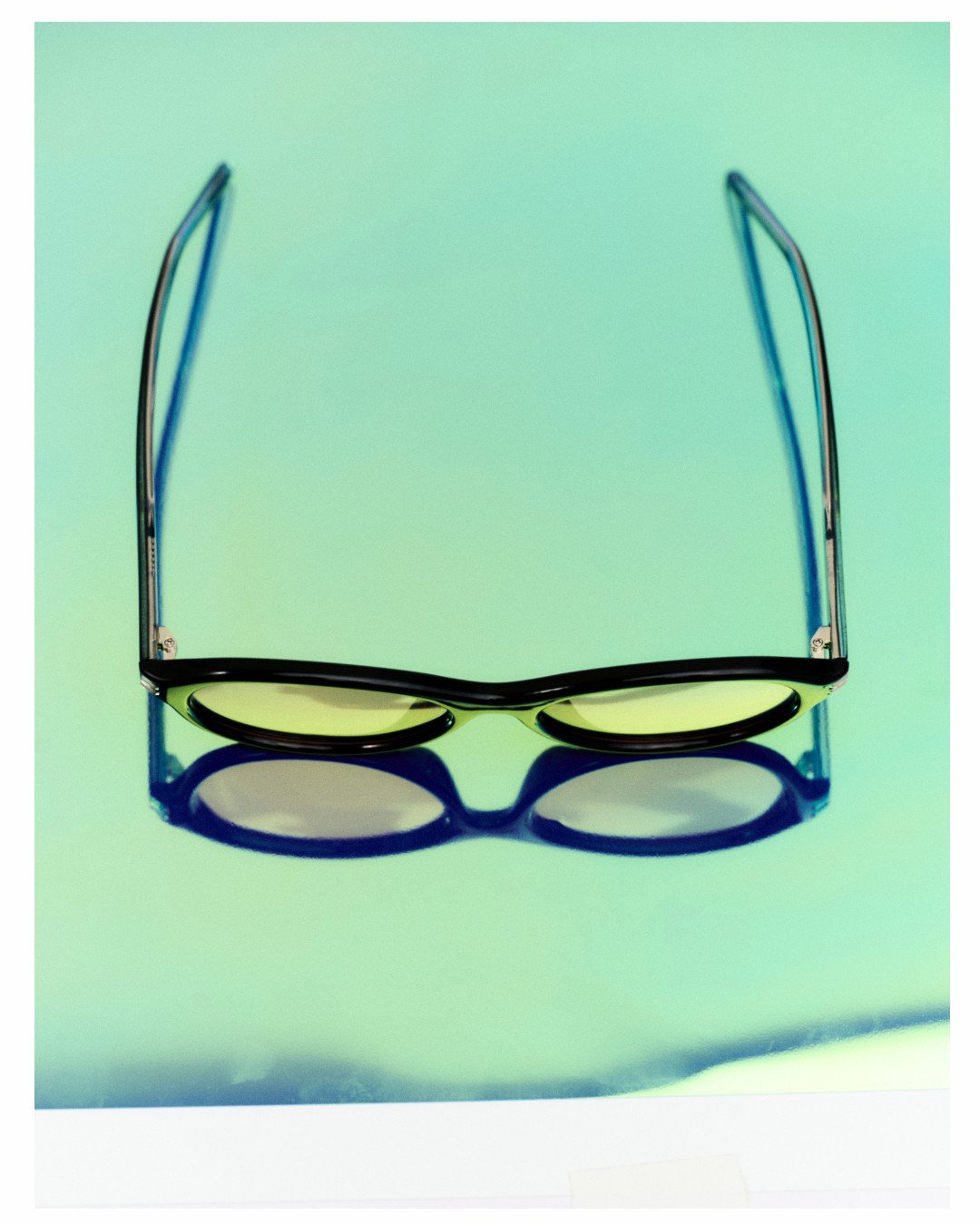 A pair of black-rimmed sunglasses with reflective yellow lenses resting on a reflective surface, casting a shadow.