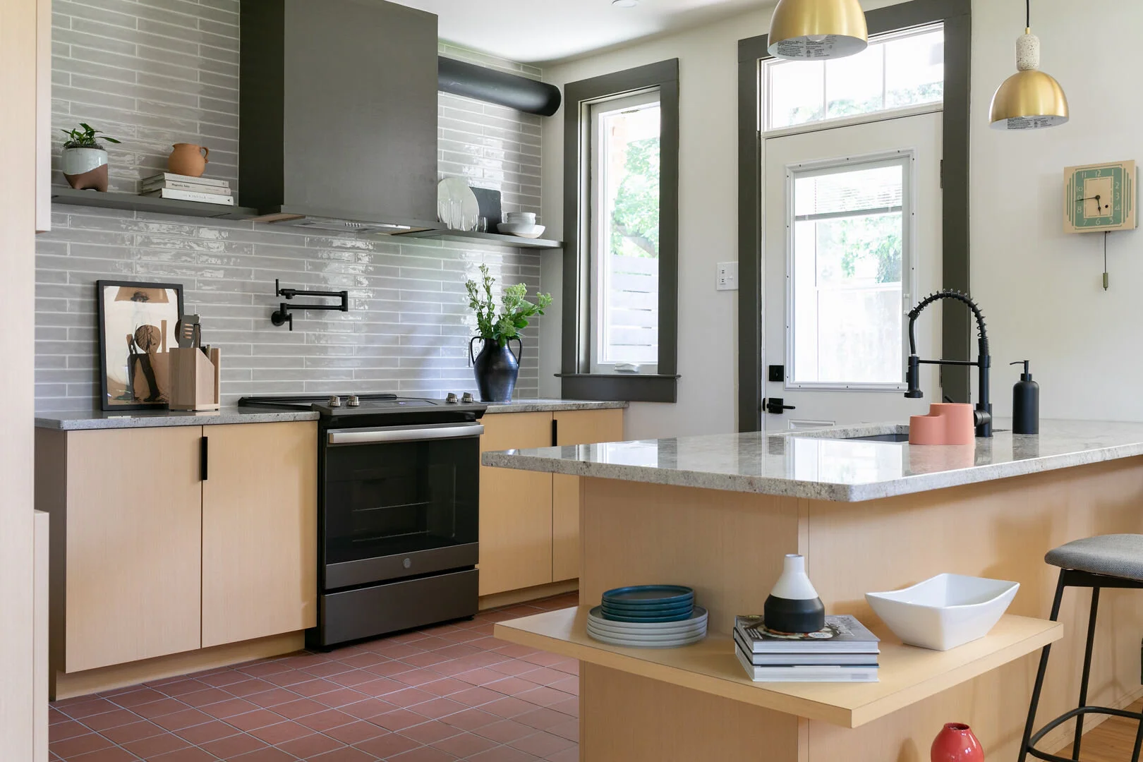 Modern kitchen with light wood cabinets, black stove, gray tile backsplash, and large windows with gray trim. Decor includes a blue vase with green foliage, stacked plates, and various small decorative objects.

Location: Lancaster, Pennsylvania