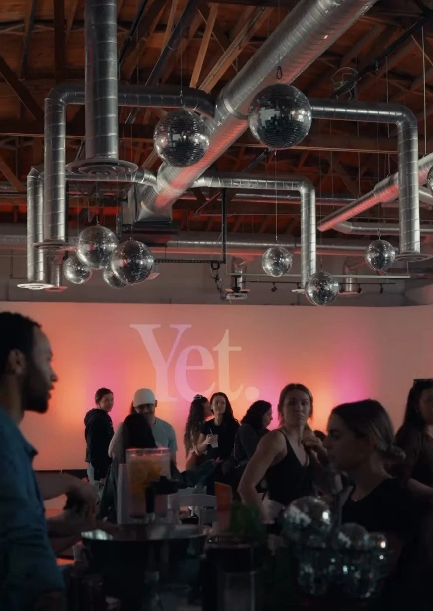 People socializing at a bar or nightclub with metallic ductwork and hanging disco balls on the ceiling, and a pink and orange gradient wall with the text 'Yet.'