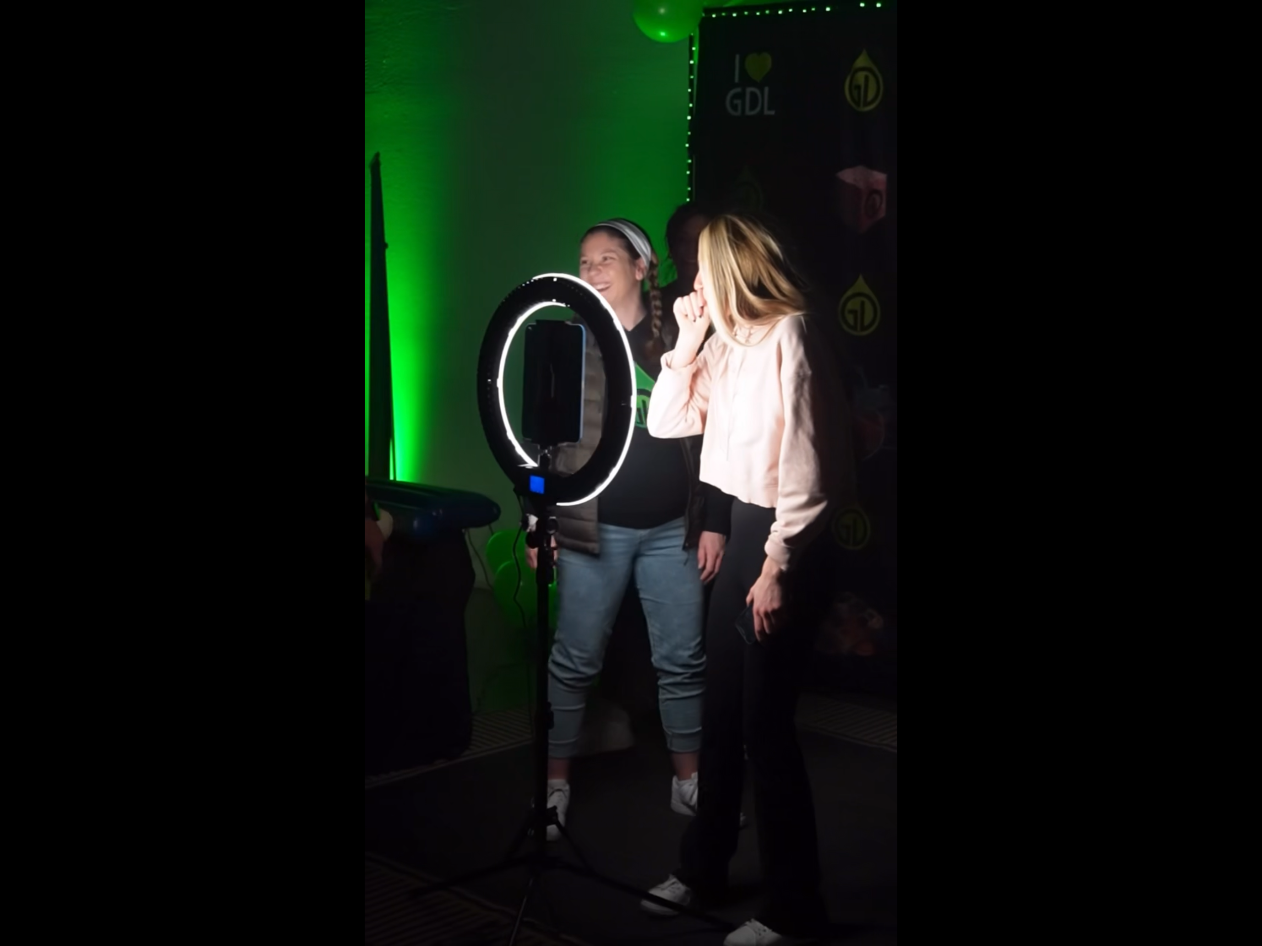 Two women looking at a smartphone mounted on a ring light, engaged in a video call or recording, in a dark room illuminated by green lights, with a backdrop displaying the phrases 'I ❤️ GDL' and various icons.