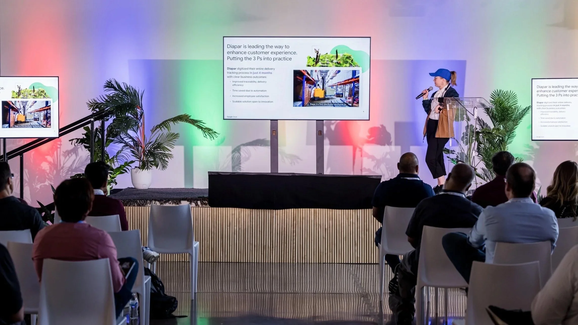 A conference room with a presenter standing at a transparent podium, speaking into a microphone. Multiple screens display presentation slides about a company called Diapaar. Attendees are seated and facing the stage, with two large plants flanking the stage. The background features a wall illuminated with colorful lights.