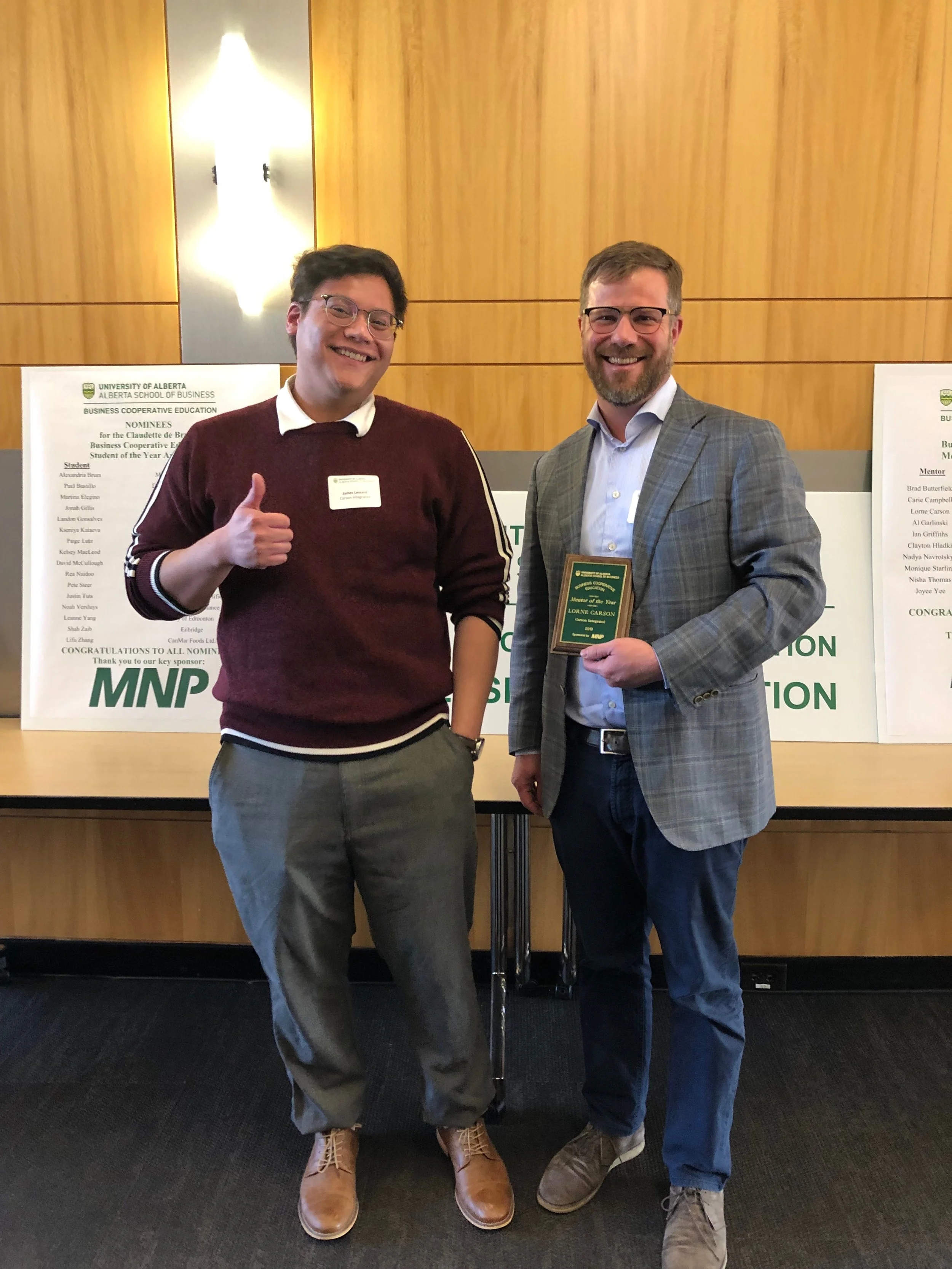 Two men stand indoors, one holding a plaque, both smiling in front of a "University of Alberta" sign, suggesting an award or recognition event.