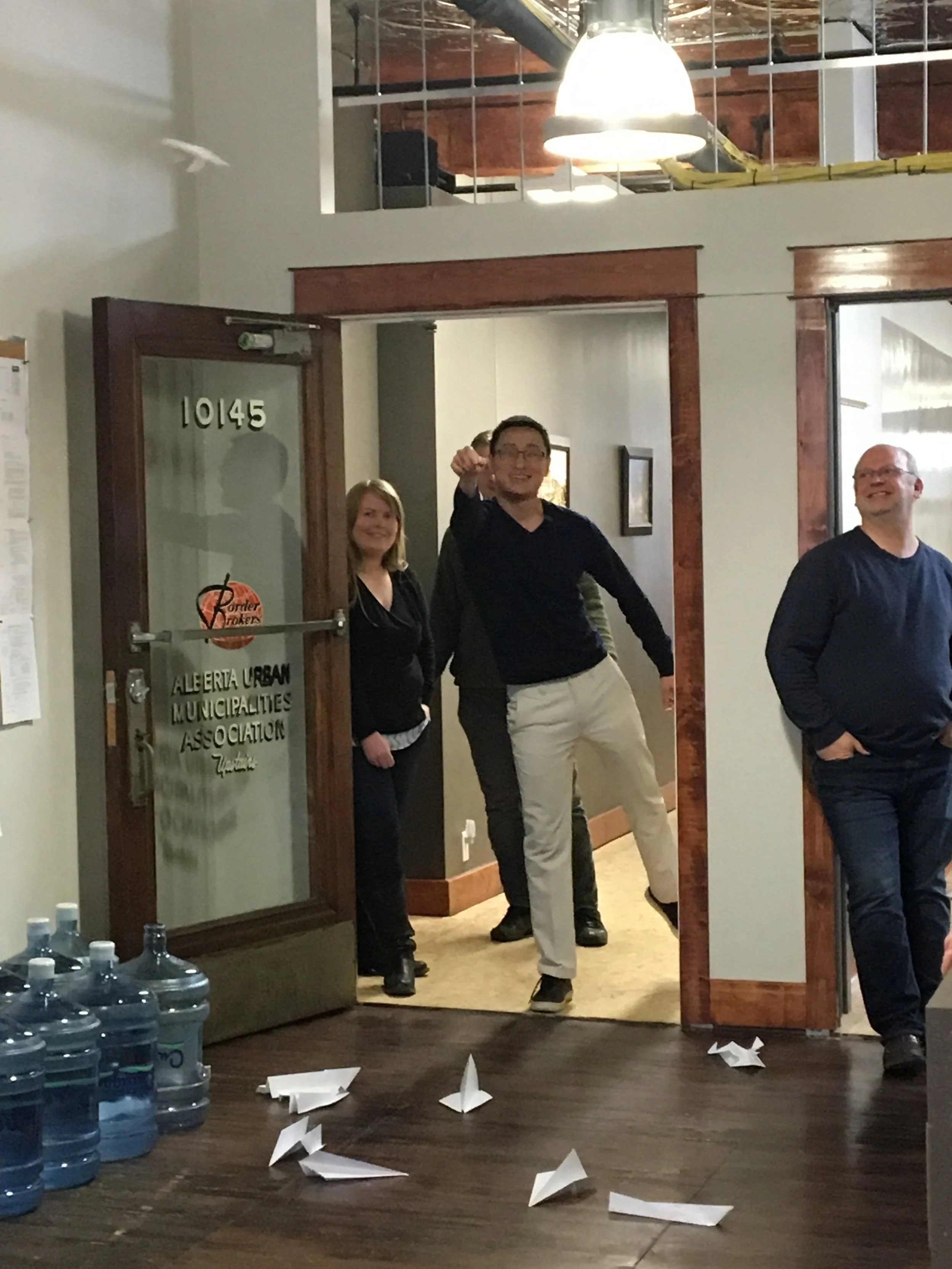 Office scene with people throwing paper airplanes, water jugs on the floor, door labeled 'Alberta Urban Municipalities Association.'
