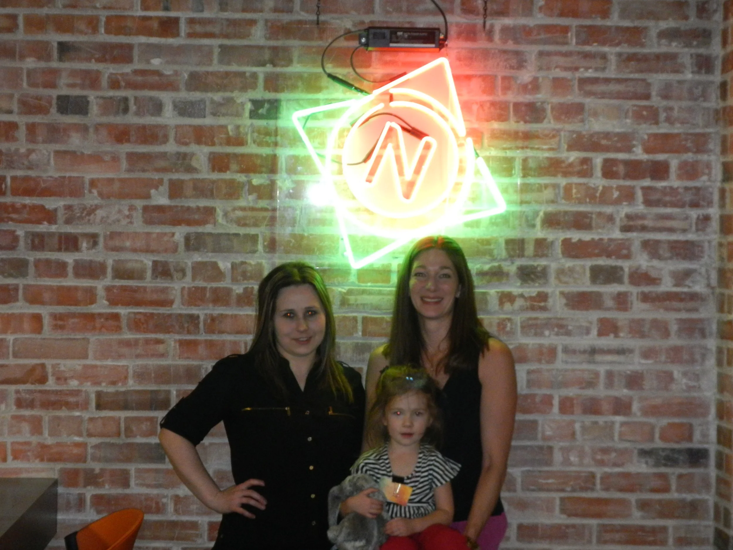 Two women and a child standing in front of a brick wall with a colorful neon sign overhead.