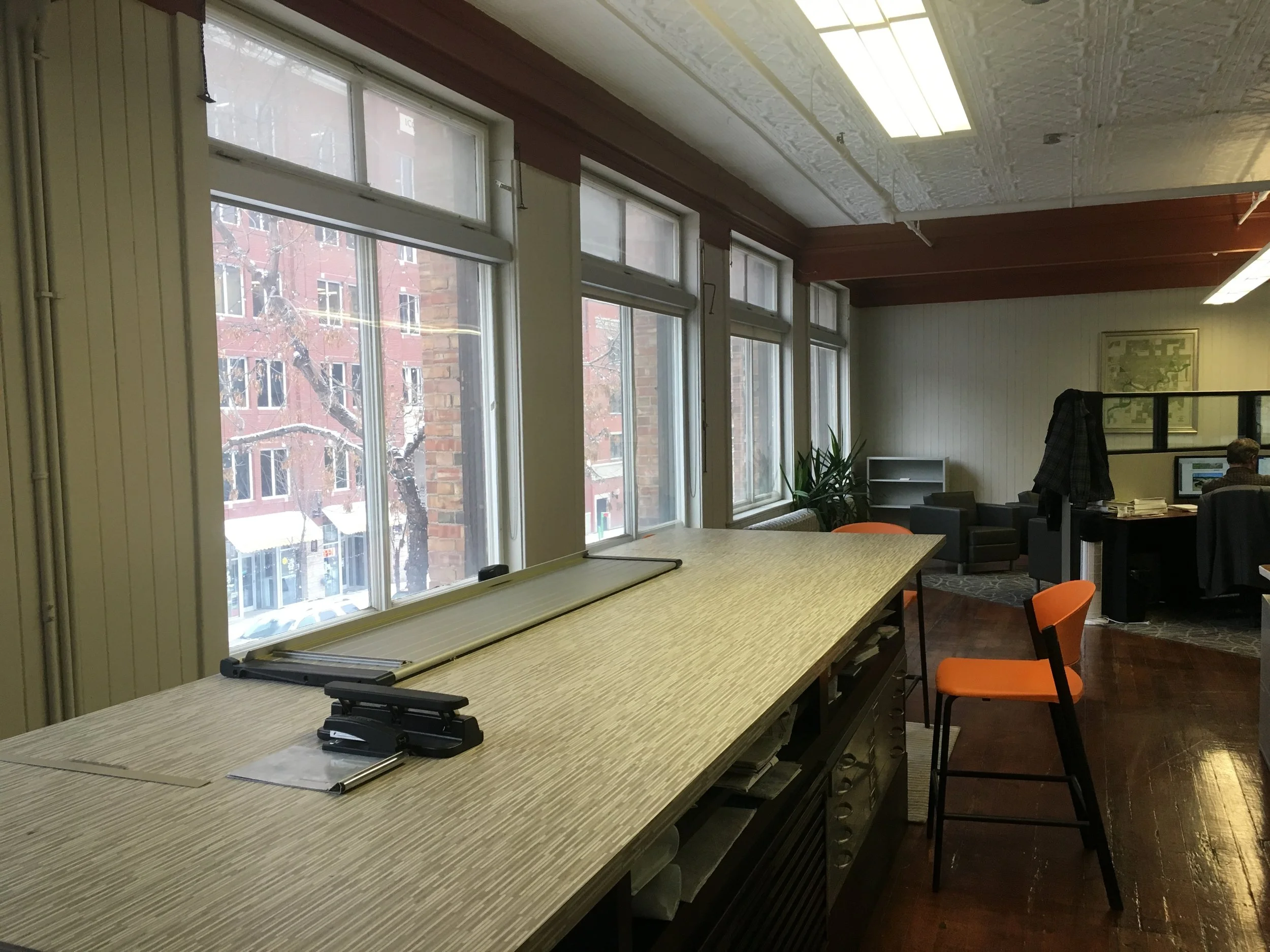 Modern office with large windows, long table, orange chairs, computer station, and plant near corner seating area.