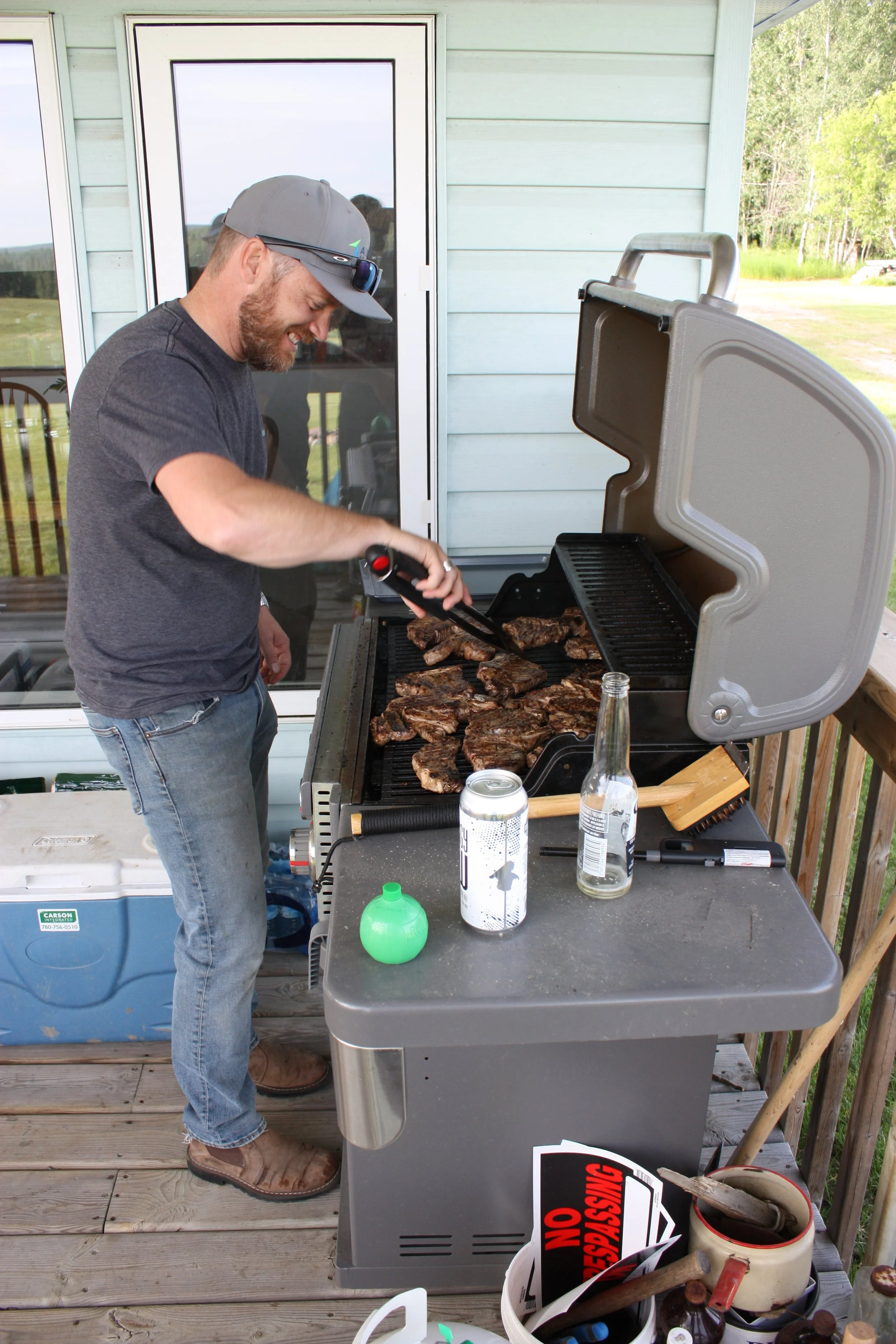 Person grilling steaks on a barbecue on a wooden deck with beverage cans nearby.