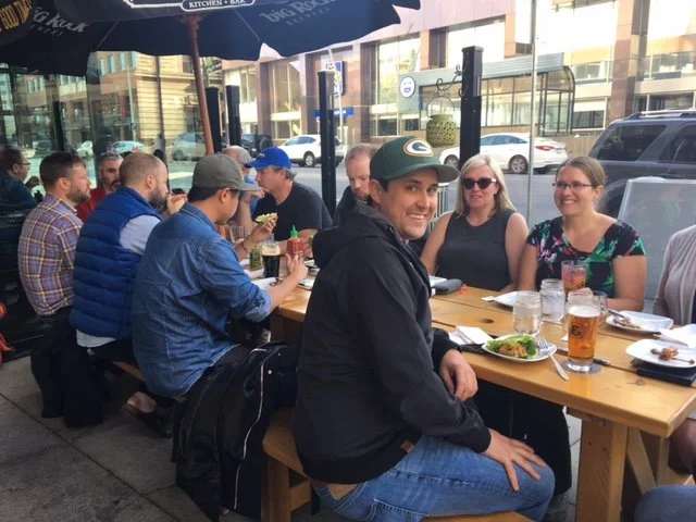 Group of people dining outdoors at a restaurant, seated around a wooden table with drinks and food.