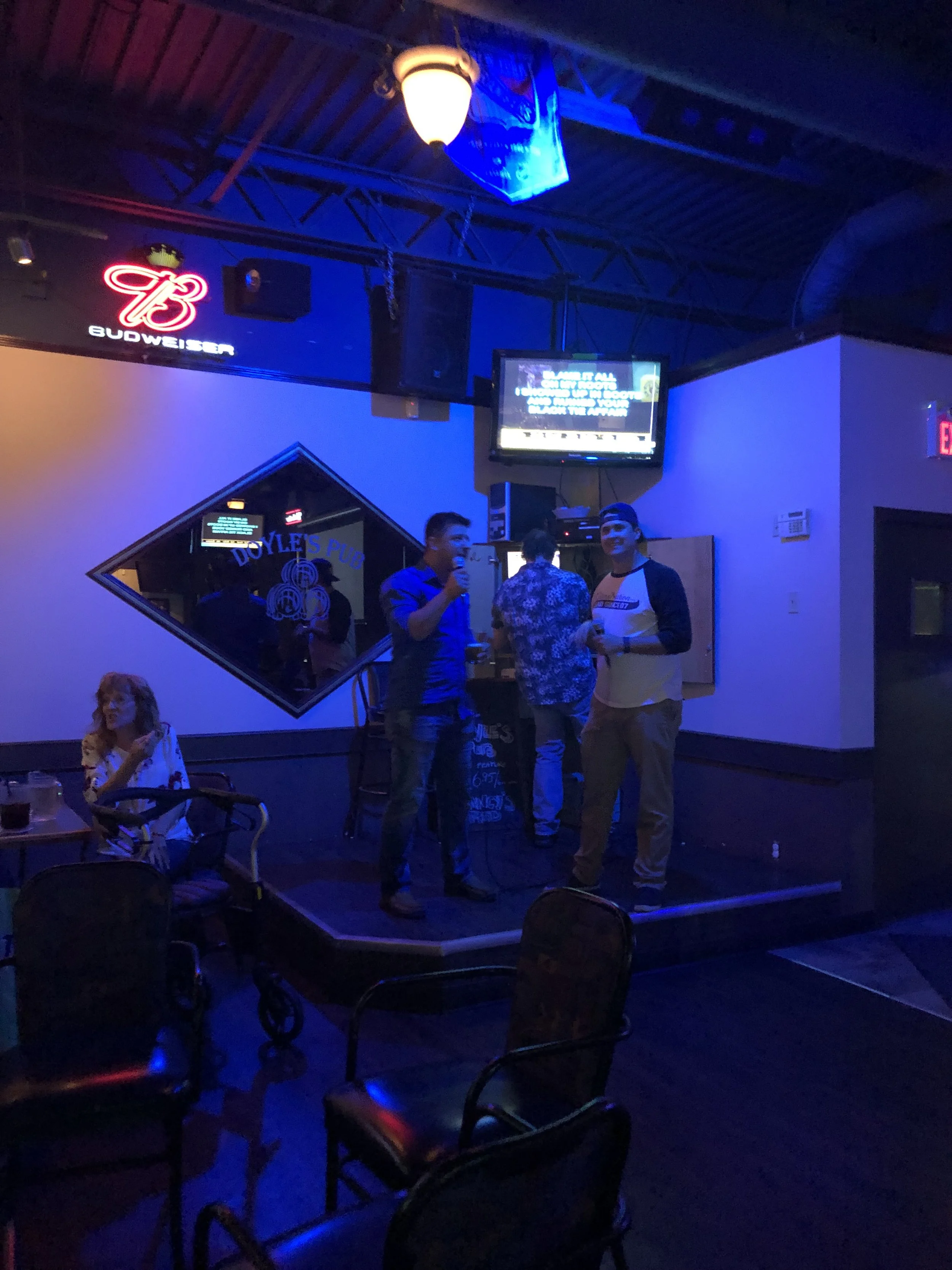 Karaoke night at a bar with patrons singing on stage and neon Budweiser sign