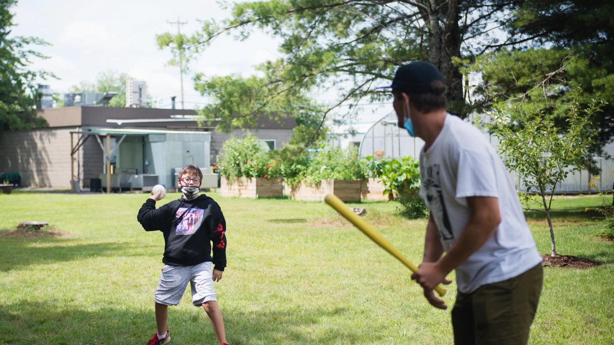 Two boys playing baseball outdoors in a backyard with trees and garden beds, both wearing masks.
