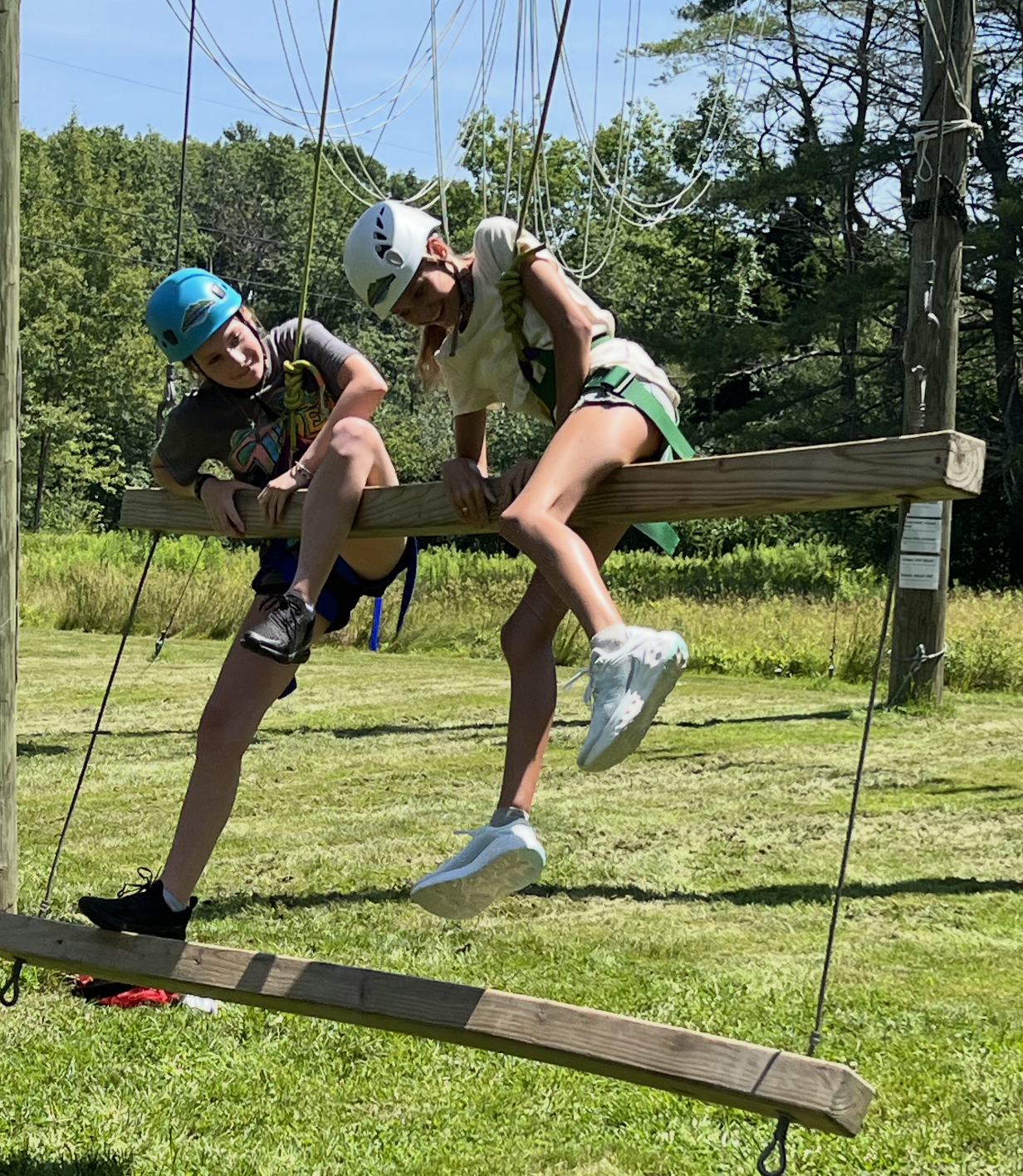 Two children on a wooden platform on ropes at an outdoor adventure park, wearing helmets and harnesses, surrounded by green trees and grass.