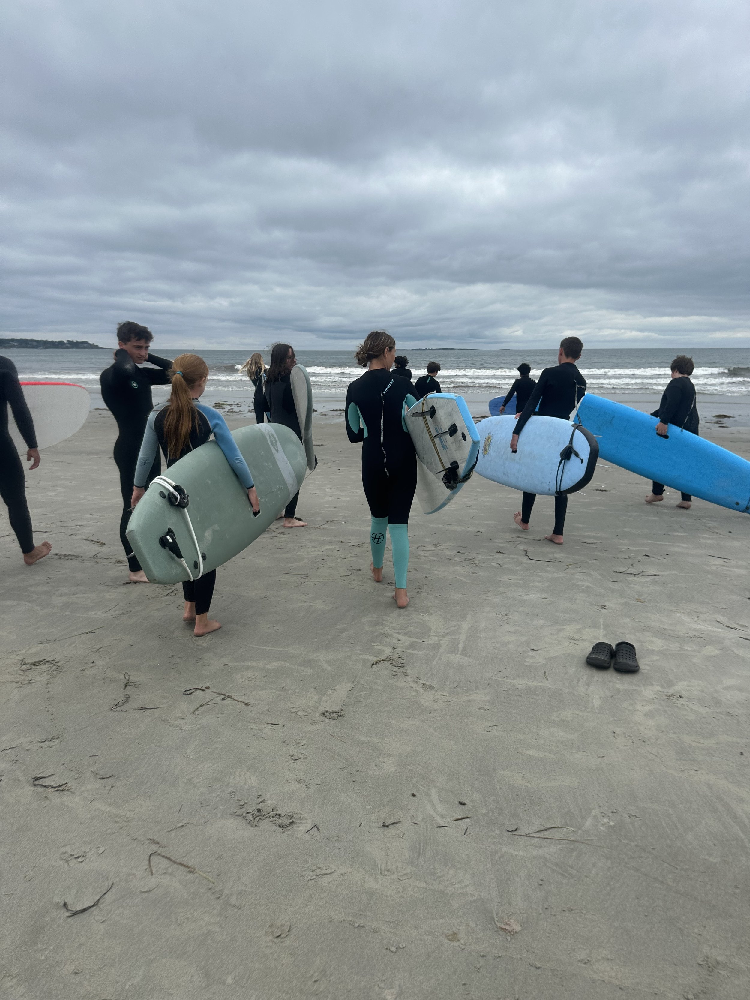 Group of young surfers walking on a sandy beach with surfboards, heading towards the ocean under cloudy skies.