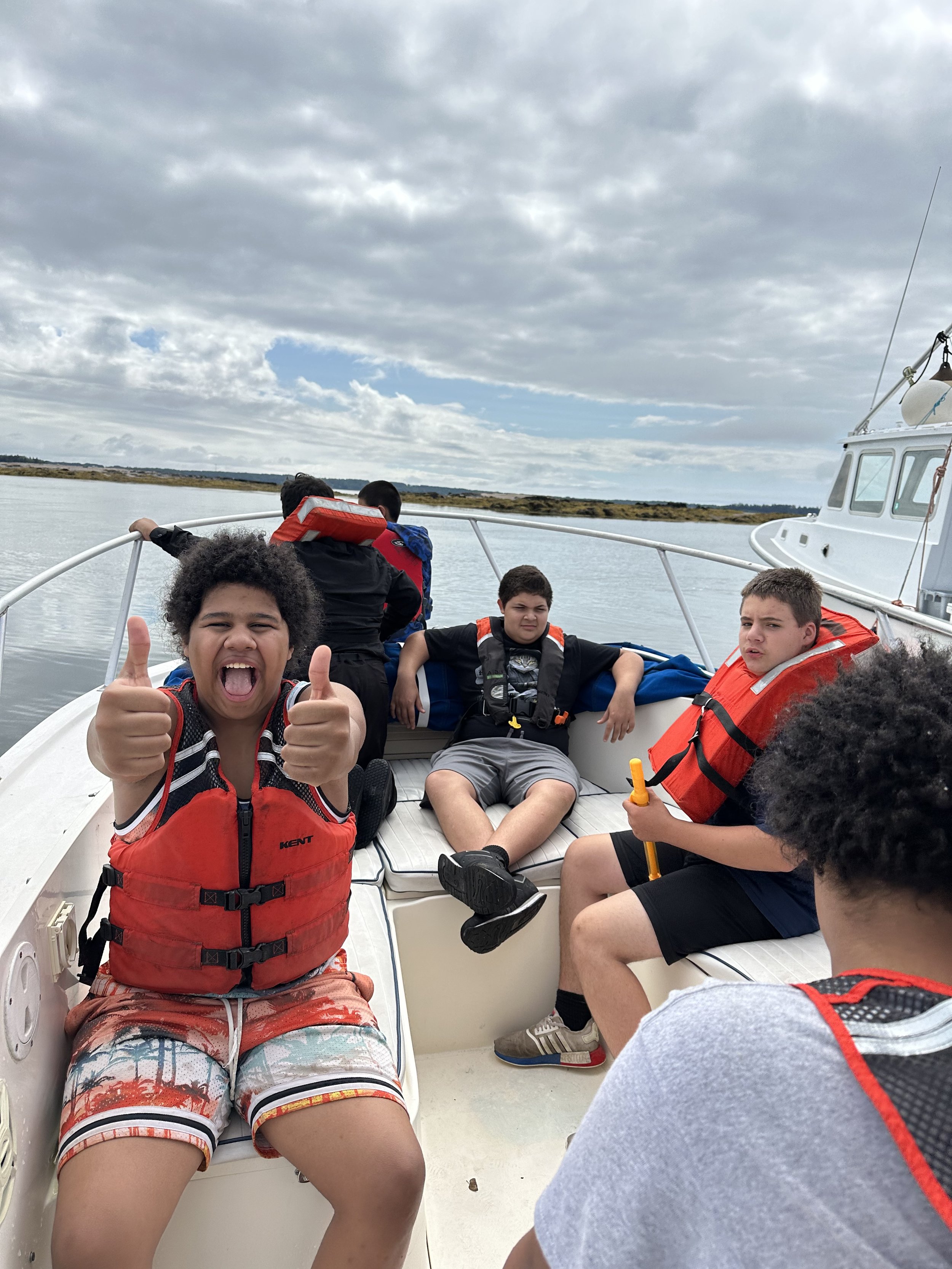 Group of children and teenagers on a boat, some wearing life jackets, enjoying a boat ride on a body of water under a cloudy sky.