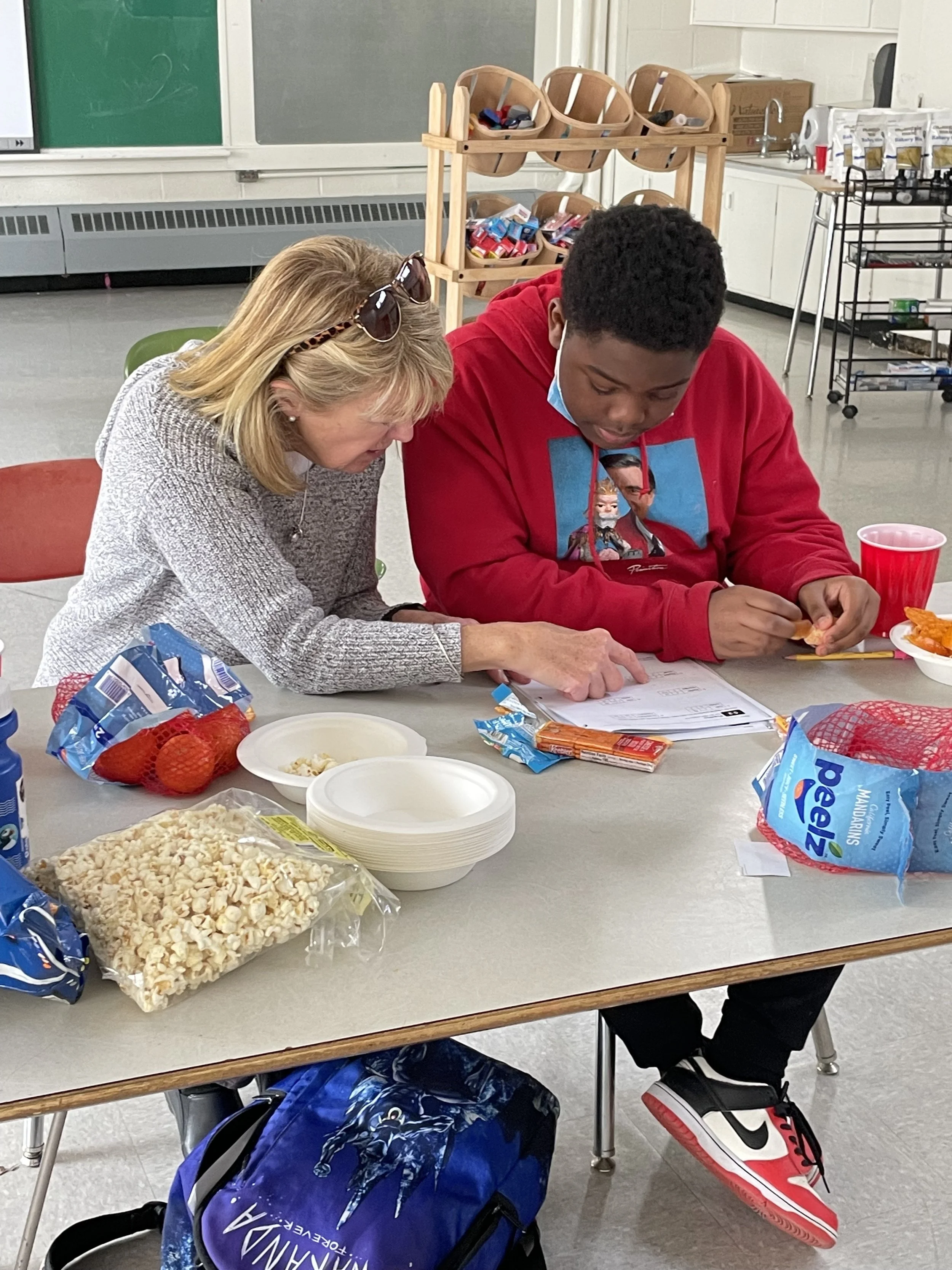 A teacher and a student sitting at a table in a classroom, looking at some papers together. The table has snacks and popcorn on it, and a backpack with the word "Wanda" is underneath. The classroom has a chalkboard, some cabinets, and school supplies in the background.