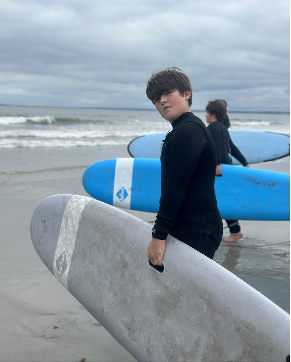 Two boys carrying surfboards on a beach, with the ocean waves and cloudy sky in the background.