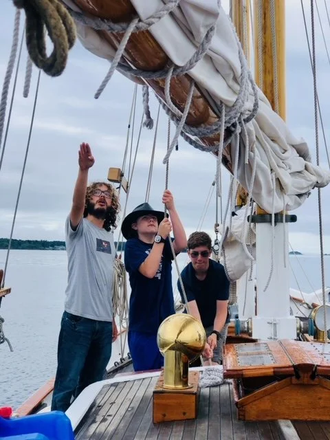 Three people on a sailboat, one pointing upwards, another holding a rope and wearing a hat, with water and land in the background.