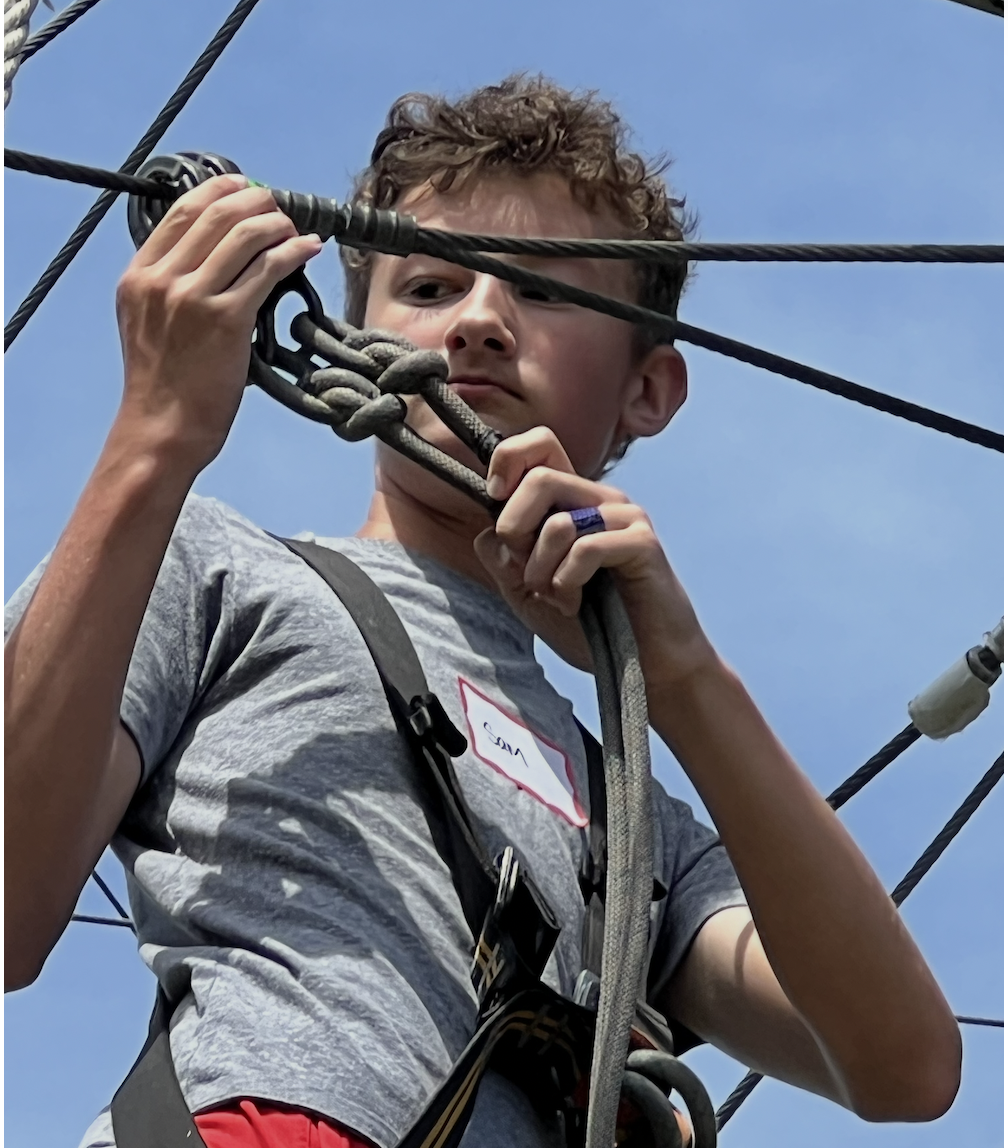 Young boy with curly hair wearing a gray shirt and harness, climbing a rope structure outdoors against a blue sky.