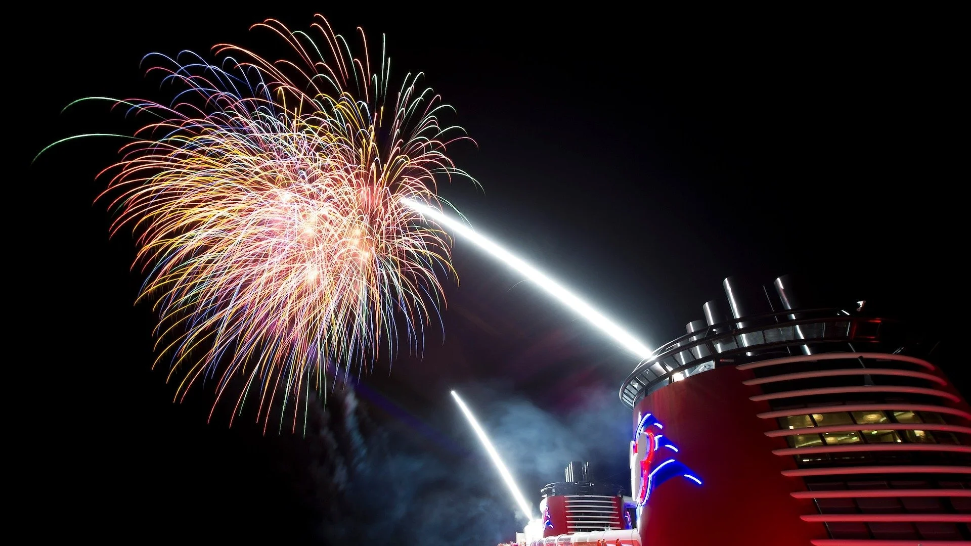 Aerial view of a busy port with five large cruise ships docked side by side along a pier, surrounded by turquoise water and a vibrant city with buildings, roads, and green areas.