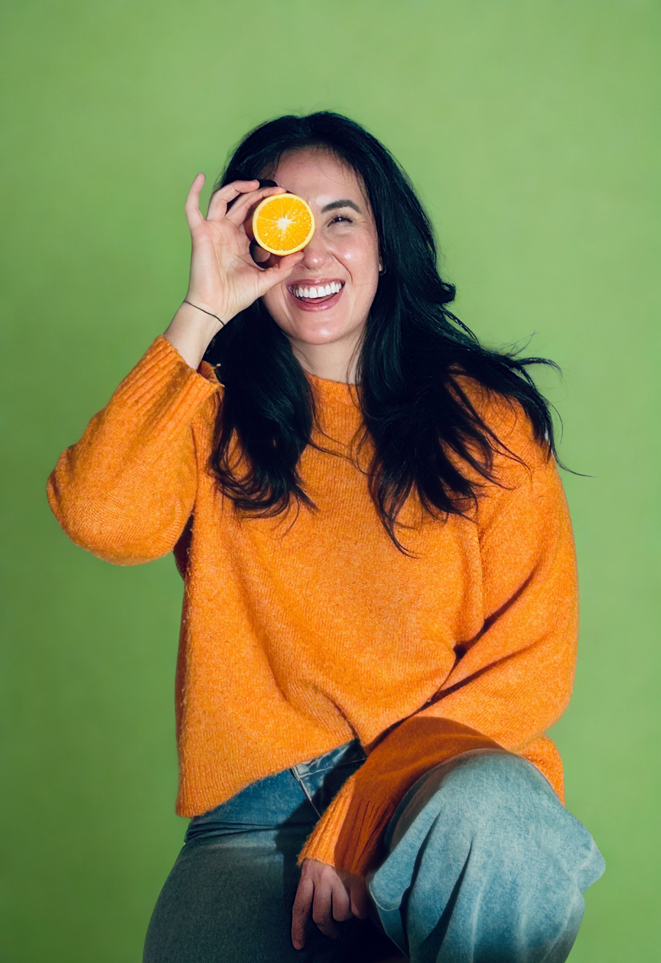 A woman with long dark hair wearing an orange sweater and blue jeans, holding a half orange up to her eye and smiling against a green background.