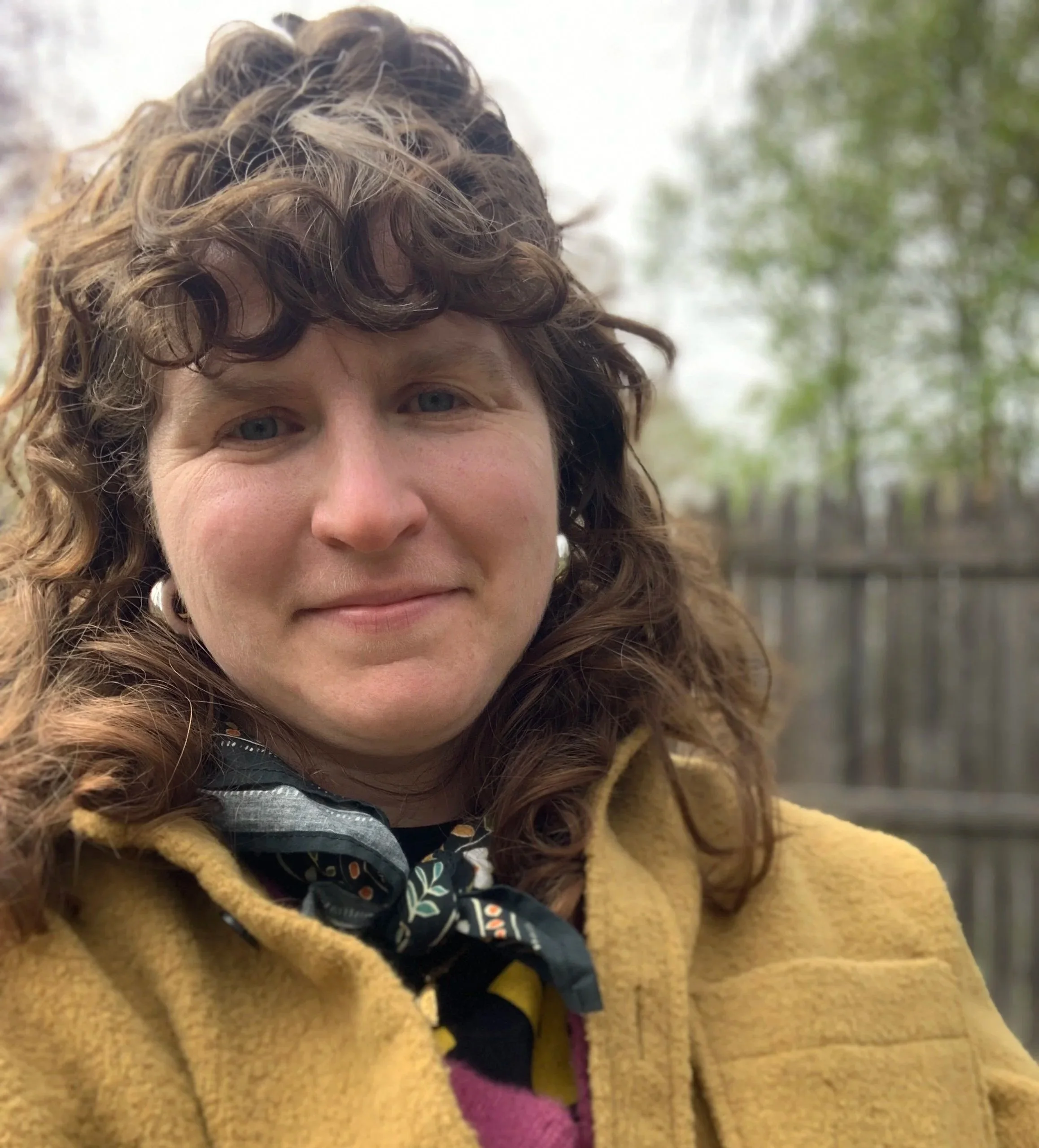 Close-up of a woman's face with curly hair, wearing a mustard yellow coat and a patterned scarf, outdoors with blurred green foliage in the background.