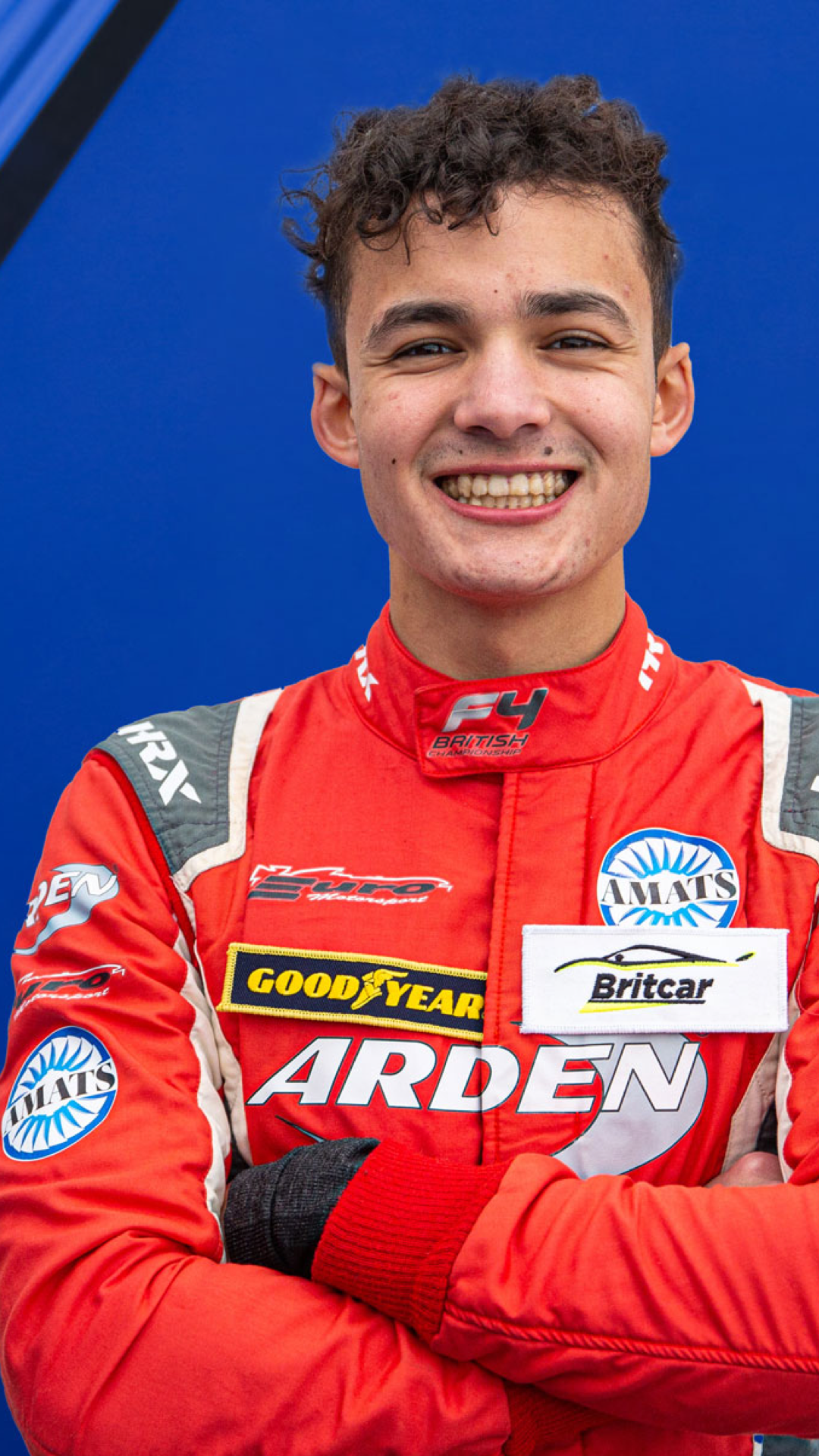 Young male race car driver wearing a red racing suit with sponsor patches, smiling with arms crossed, standing in front of a blue background.