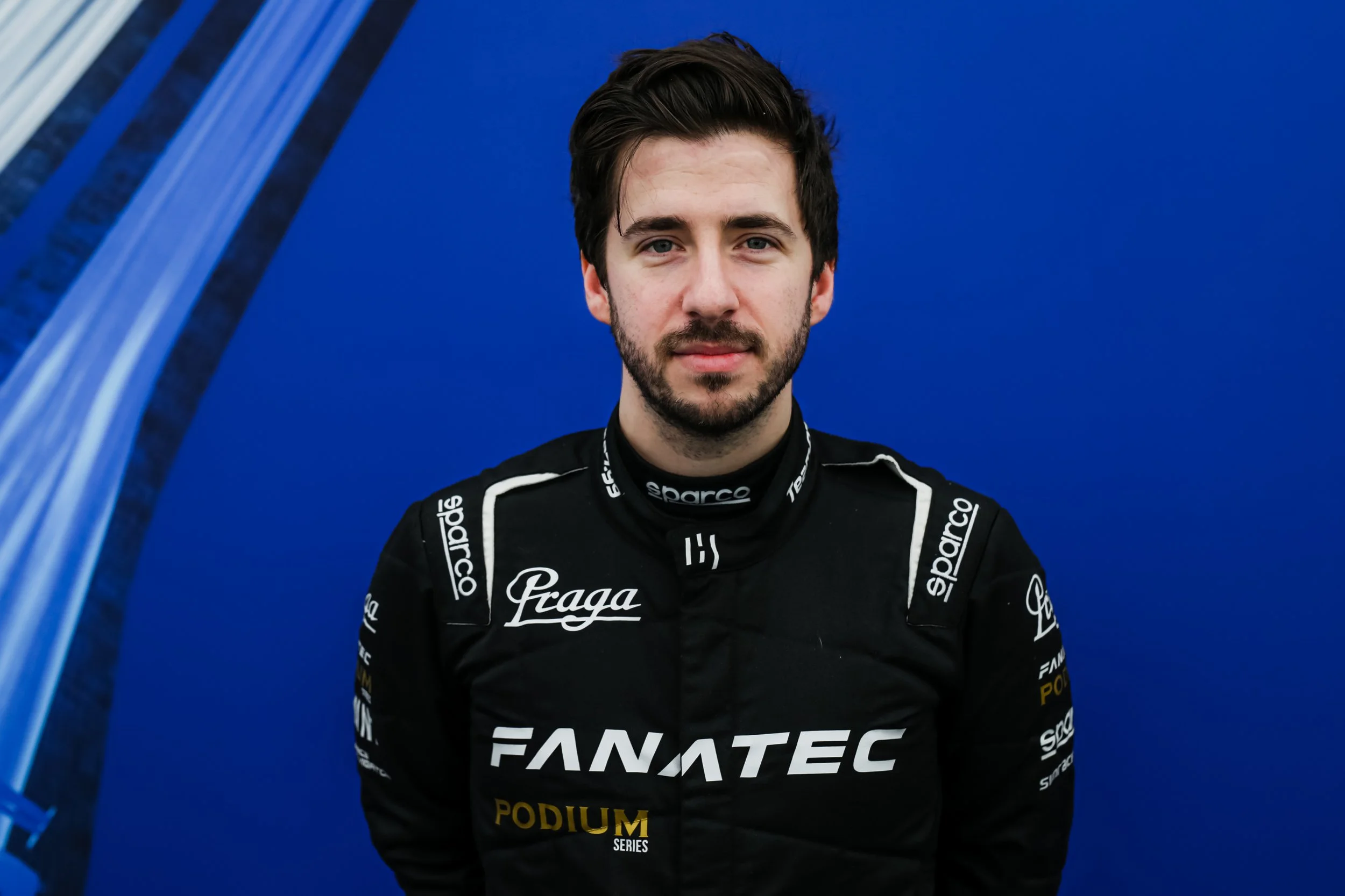 Jimmy Broadbent in a black racing suit with sponsor logos, standing in front of a blue background.