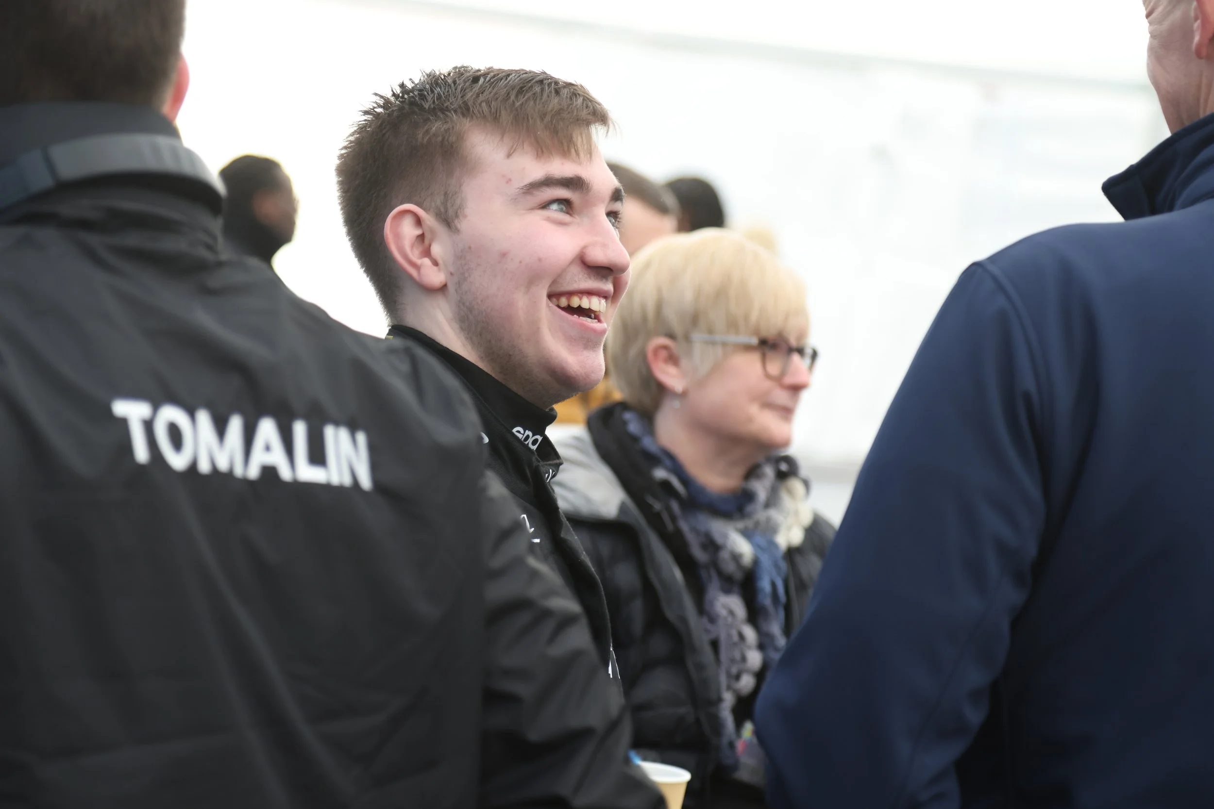 Gordie Mutch smiling and talking to people at an event, wearing a black racing suit.
