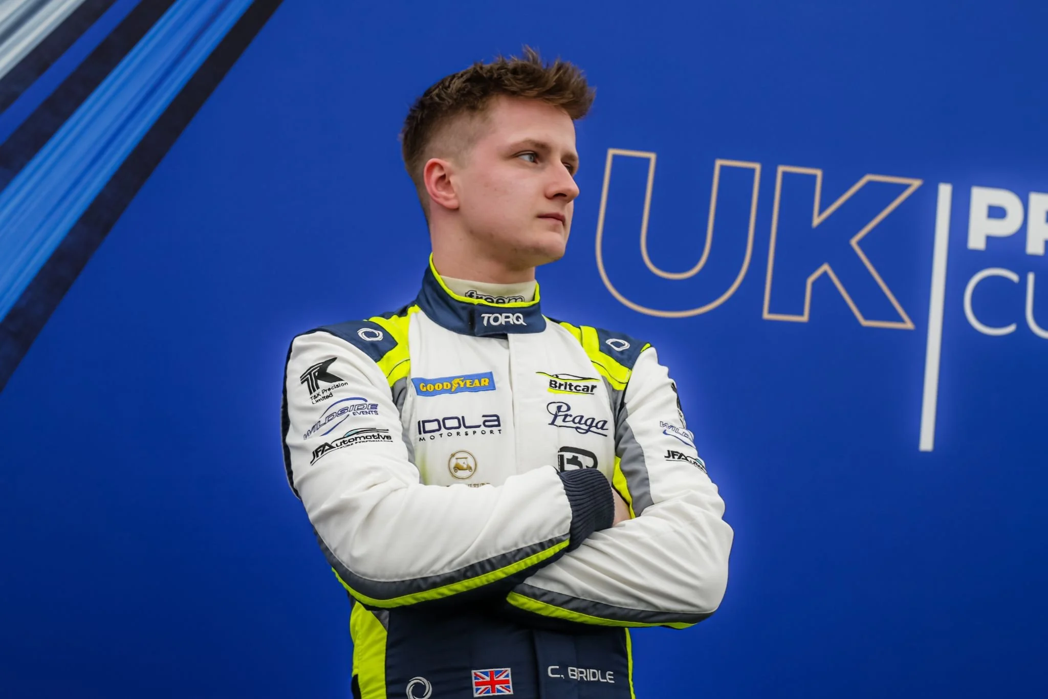 Chris Bridle - Young male race car driver standing with arms crossed, wearing a racing suit with various sponsor patches, in front of a blue background.