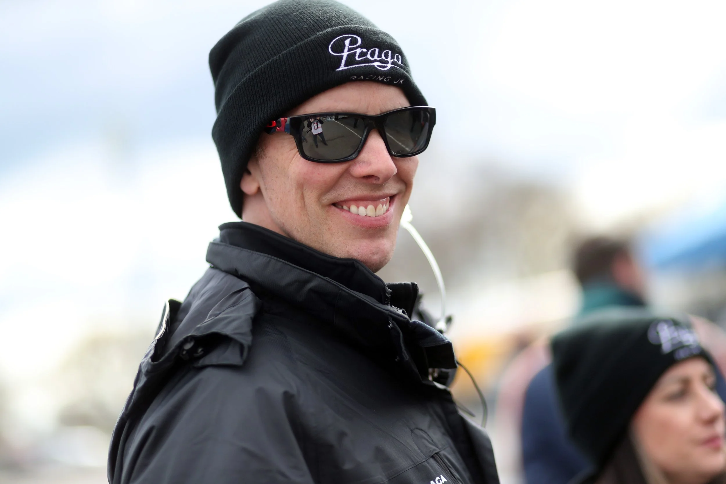 Al Boulton smiling wearing sunglasses, a black beanie with 'Praga' embroidered on it, and a black jacket, standing outdoors.