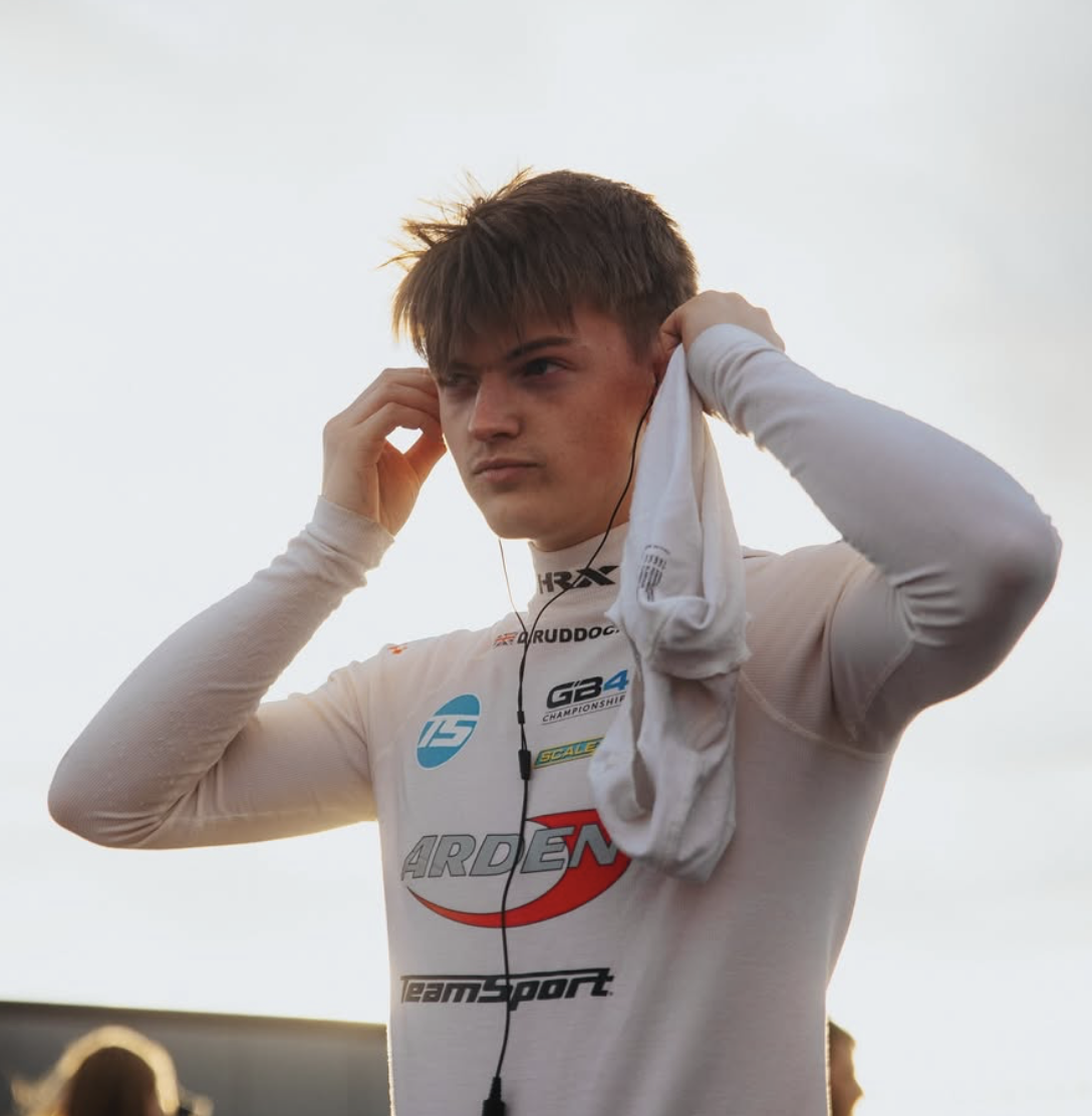 Young male race car driver with short brown hair, in a white racing suit with various sponsor logos, adjusting headphones outdoors with a cloudy sky background.