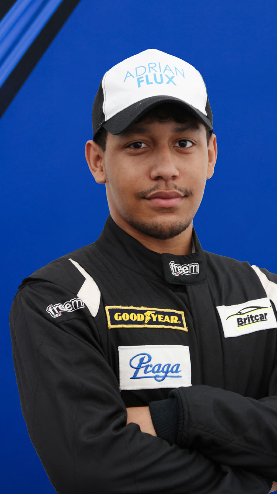 Young race car driver wearing a black racing suit with sponsor patches, crossed arms, and a white and black cap with "Adrian Flux" written on it, standing against a blue background.