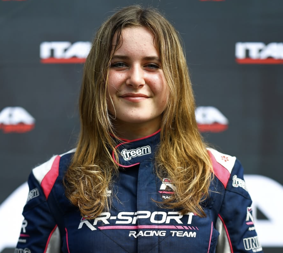 Young female race car driver wearing a racing suit with team logos, smiling against a backdrop with branding.