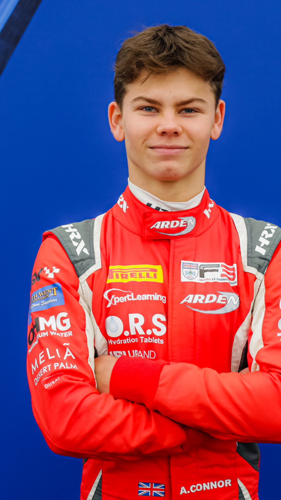 Young male racing driver in red racing suit with sponsor logos, arms crossed, standing against a blue background.