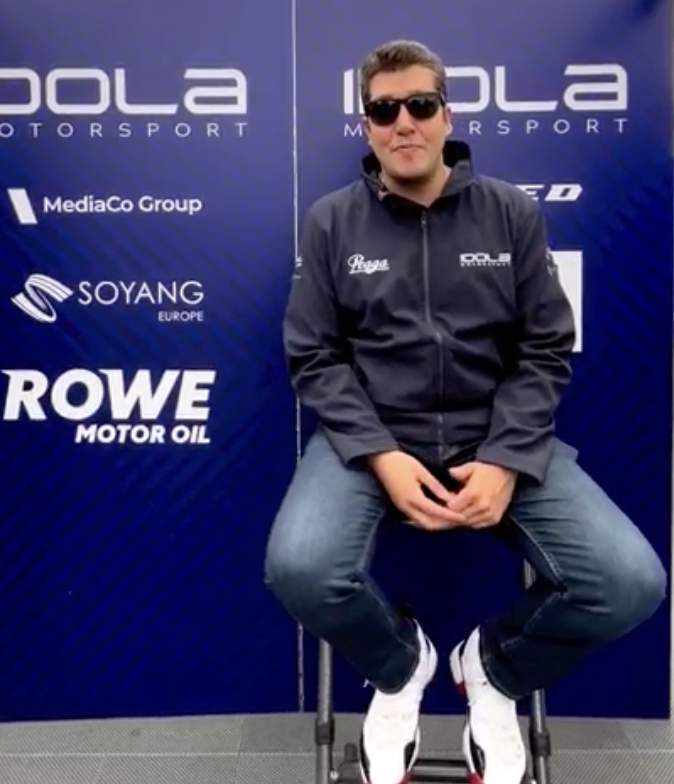 Rodolfo Gonzalez sitting on a stool in front of a blue backdrop with racing and automotive sponsor logos, wearing sunglasses, a dark jacket, jeans, and white sneakers.
