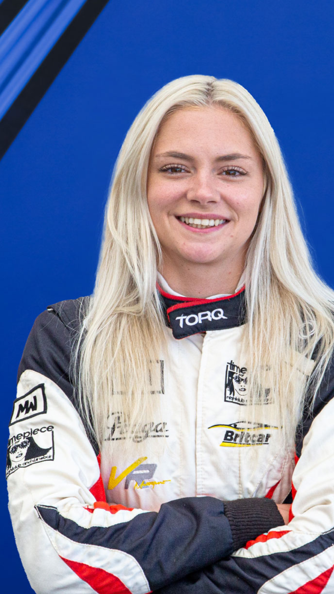 A female race car driver in a racing suit with various sponsor logos, smiling with arms crossed, against a blue background.