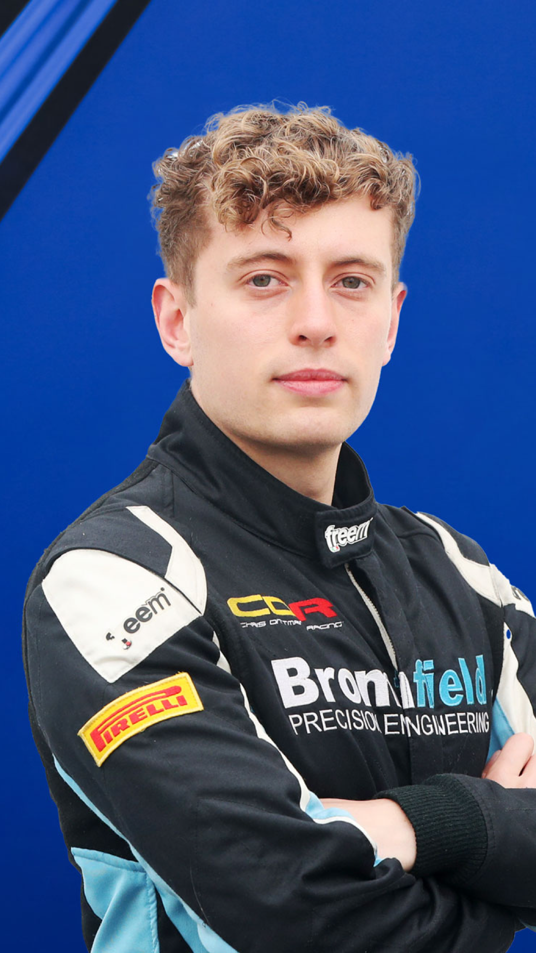 Alex Fores - Race car driver in a racing suit with arms crossed, standing against a blue background.