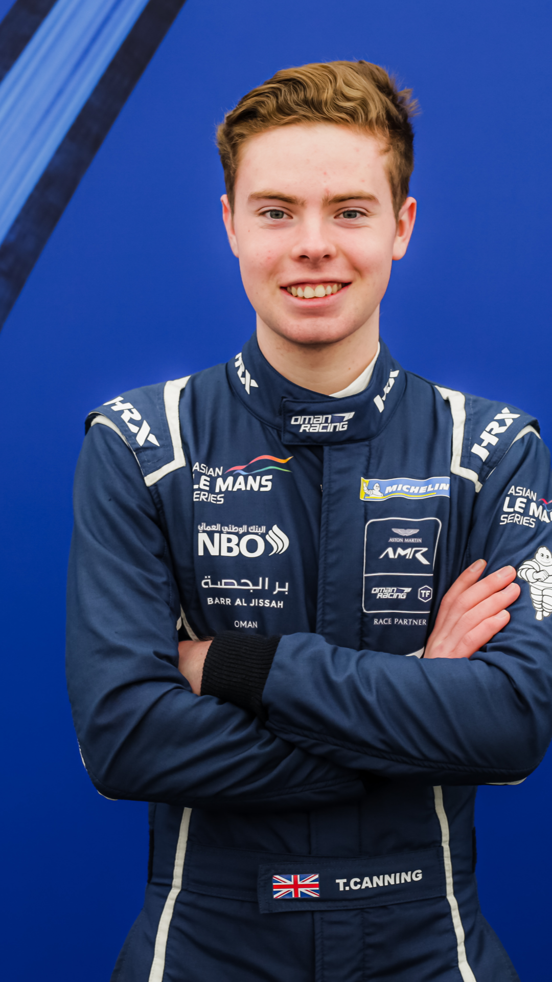 Tom Canning in blue racing suit with sponsor logos, arms crossed, posing in front of a blue background.