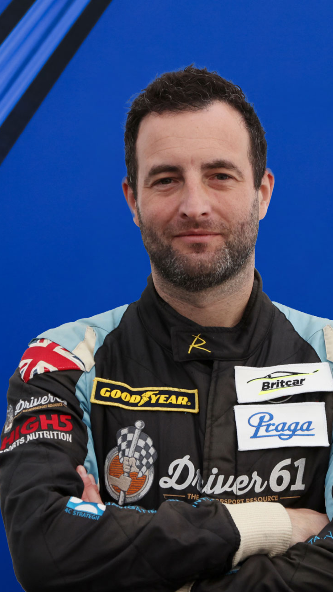 A race car driver in a racing suit standing with arms crossed, against a blue background.