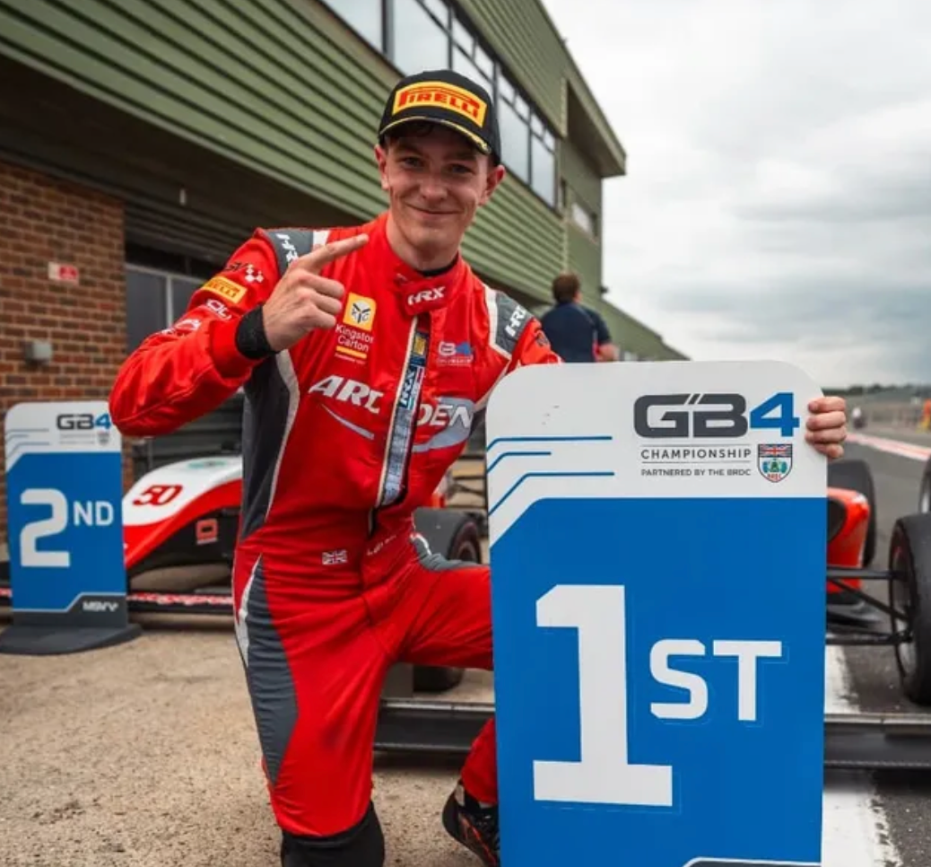 A race car driver in a red racing suit kneeling next to a sign that reads 1st place, with a race car and another second place sign in the background. The driver is smiling and pointing at himself, wearing a Pirelli cap.