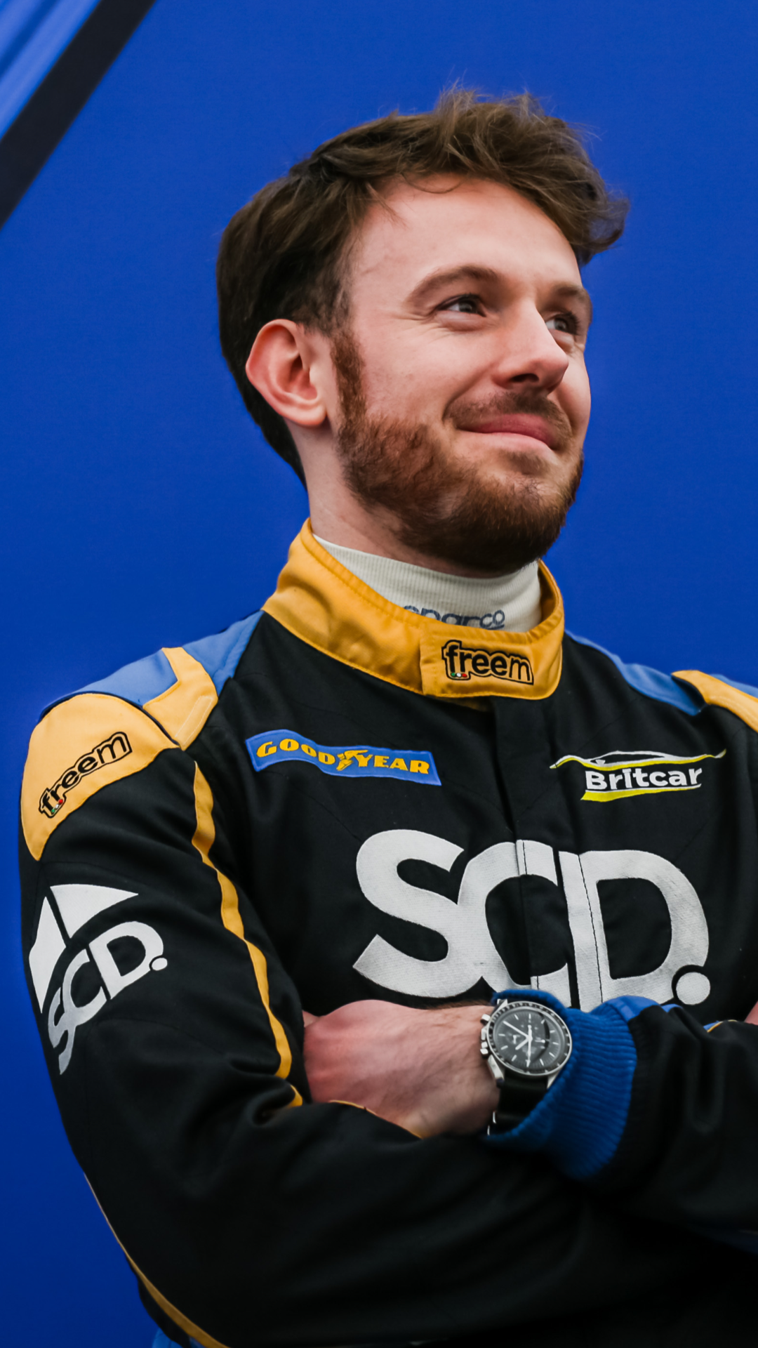 Miles Lacey - A race car driver in a black and yellow racing suit with sponsor logos, standing with arms crossed and smiling, against a blue background.