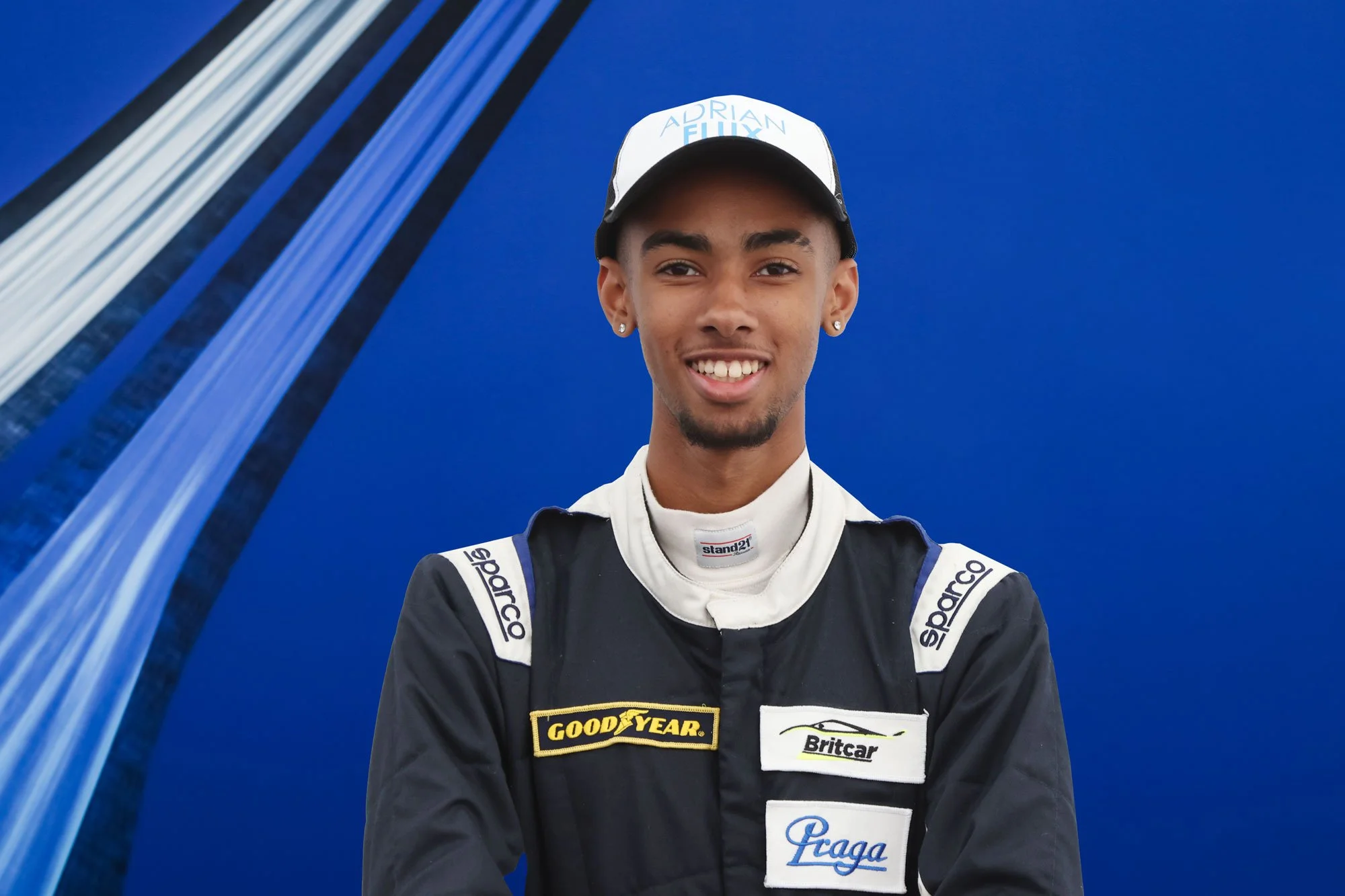 Young race car driver wearing a black racing suit with sponsor patches, smiling at the camera, against a blue background.