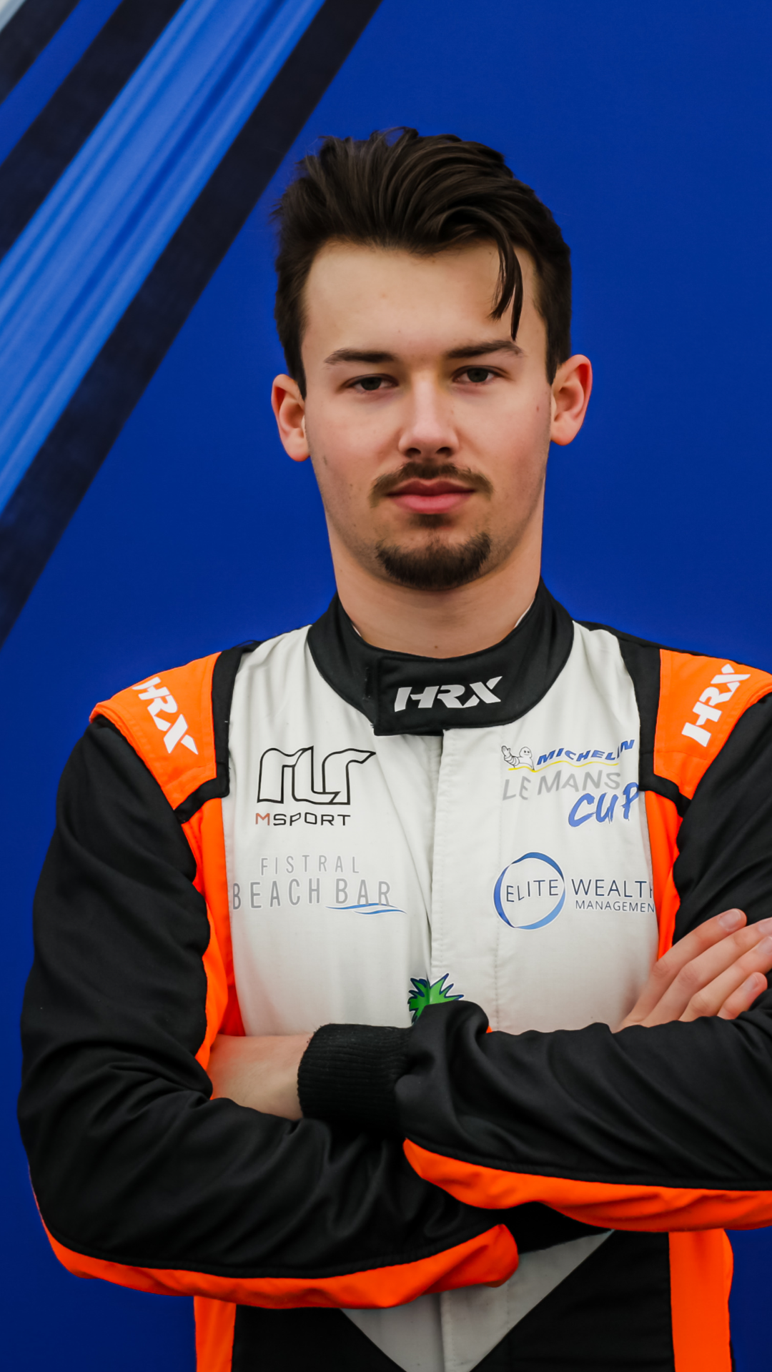 Tommy Foster - A male race car driver wearing a white and black racing suit with orange and black detailing, standing with crossed arms against a blue background.