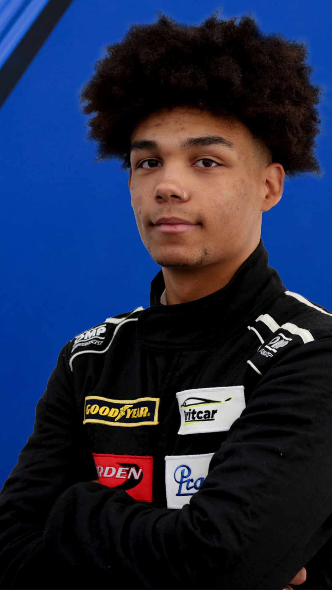 Young man in a racing suit with sponsor patches, standing against a blue background.