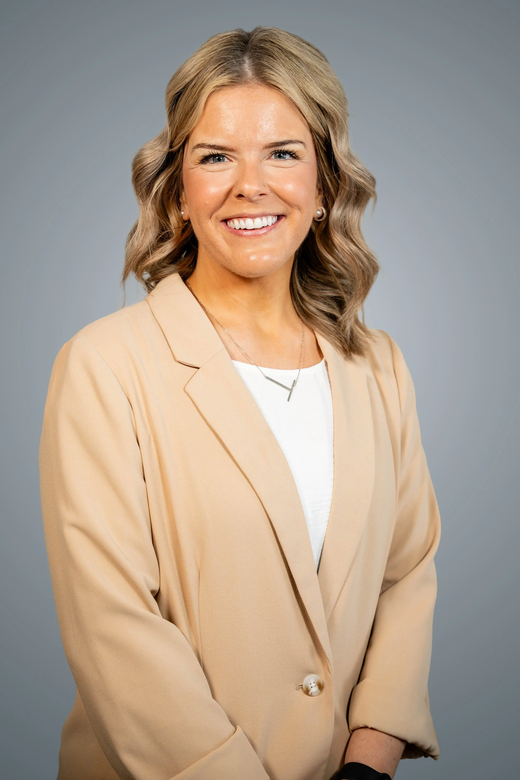 Portrait of a smiling woman with shoulder-length blond hair, wearing a maroon top, hoop earrings, pearl earrings, and a silver necklace with a cross pendant.