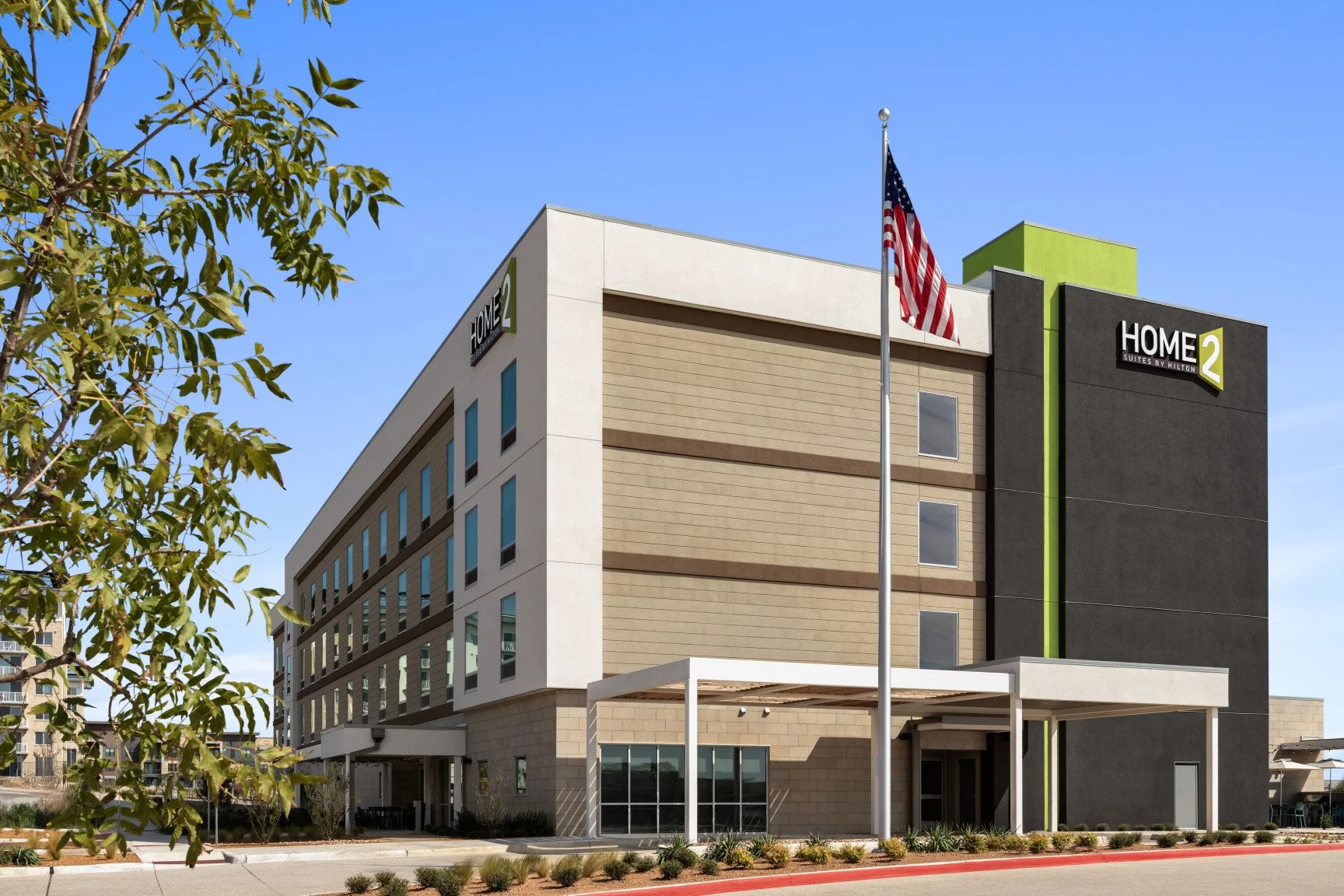 Exterior of a modern hotel building with a sign that reads 'Home2 Suites by Hilton'. American flag on a flagpole in front of the hotel, blue sky overhead.