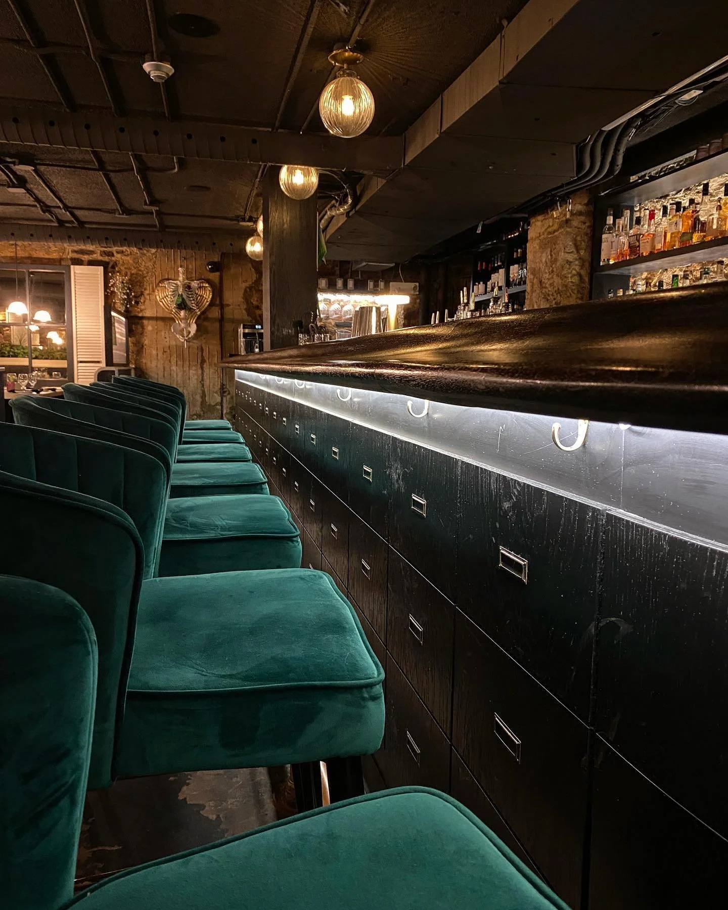 Interior of a dimly lit bar with green velvet chairs lined up at a dark wooden counter, shelves with liquor bottles, and hanging globe lights. emerald velvet booths, a high end setting in a heritage building, were past and present meet