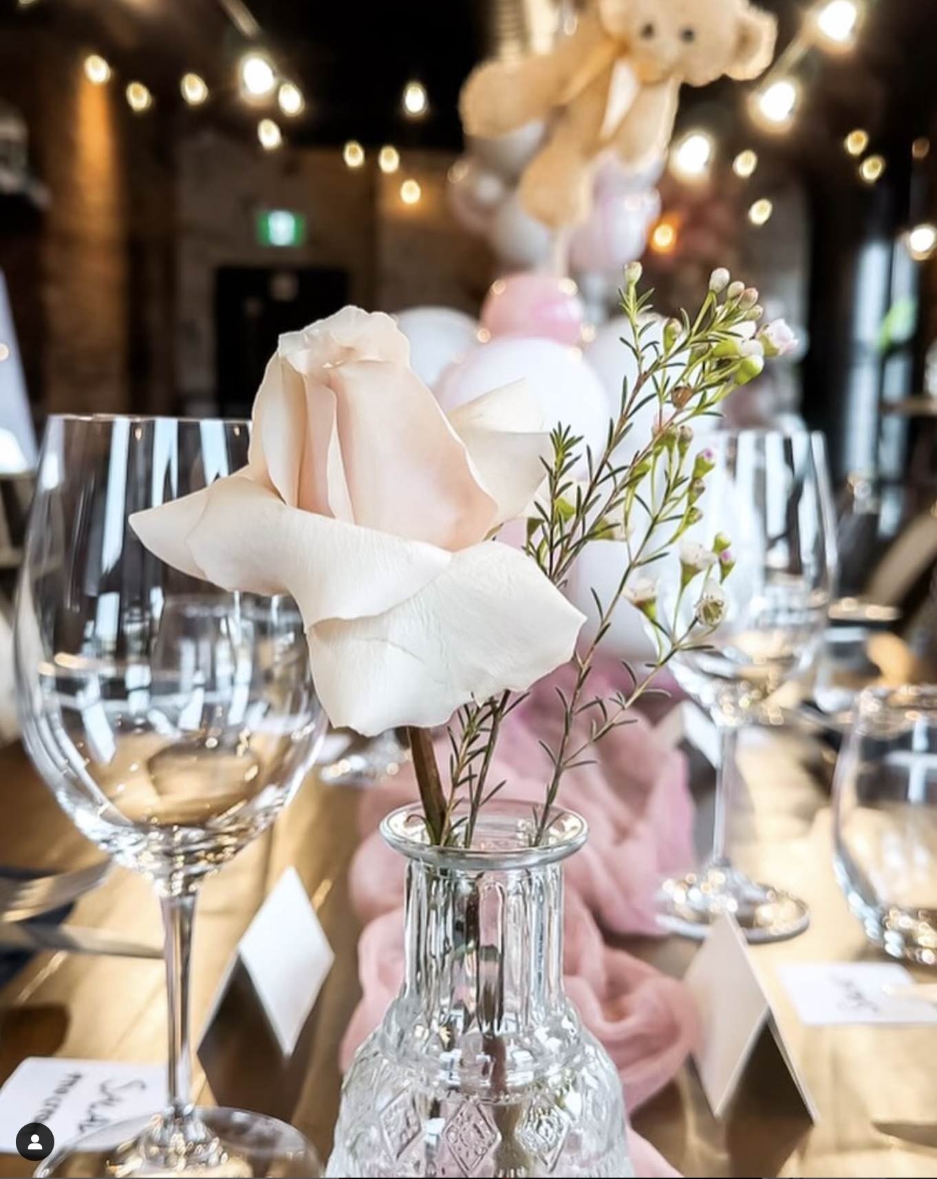 A close-up of a floral centerpiece with a single pink rose and small white flowers in a clear glass vase, set on a dining table with empty wine glasses in the background, illuminated by warm string lights in a rustic indoor setting.